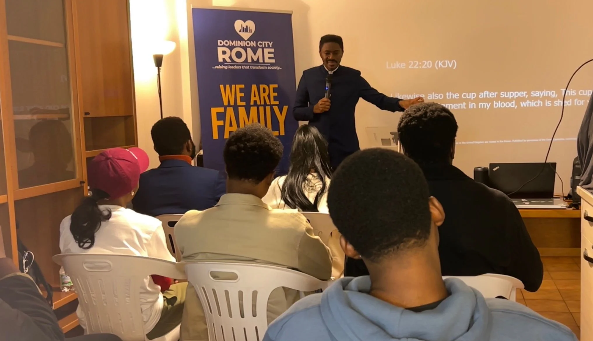 A man in a dark suit giving a presentation to an indoor audience with a blue banner that reads 'Dominion City ROME' and 'We Are Family' in orange. The screen behind him displays a Bible verse, Luke 22:20 (KJV), about the cup after supper and blood.
