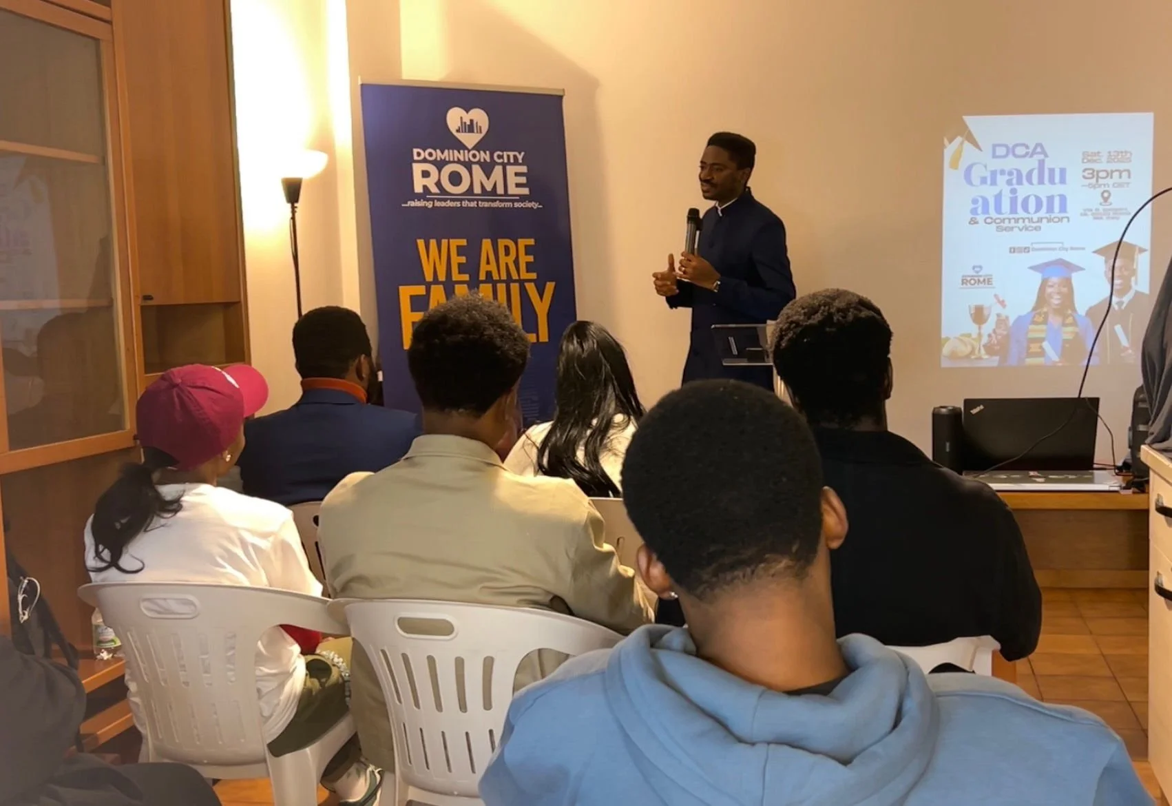 A man giving a presentation to an audience of diverse individuals in a room. There is a blue banner with the words 'Dominion City Rome' and the phrase 'We Are Family.' A projection screen shows an event poster titled 'Graduation & Communion Service,'