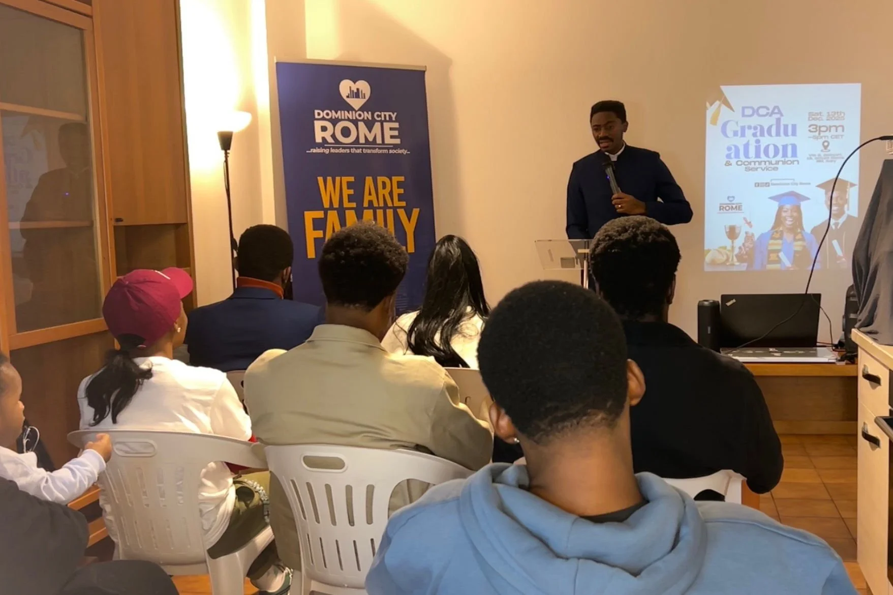 A man standing at a podium speaking to an audience during a graduation event held indoors. The audience is seated on white plastic chairs, and there is a blue banner on the left that reads 'Dominion City Rome, We Are Family.' A projected slide on the