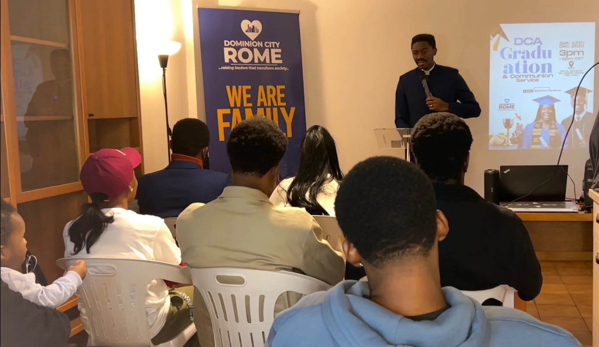 A young man in a dark suit speaking at an indoor event with an audience seated in white chairs, a blue banner that reads 'We Are Family' and a projected slide about graduation and communication services.