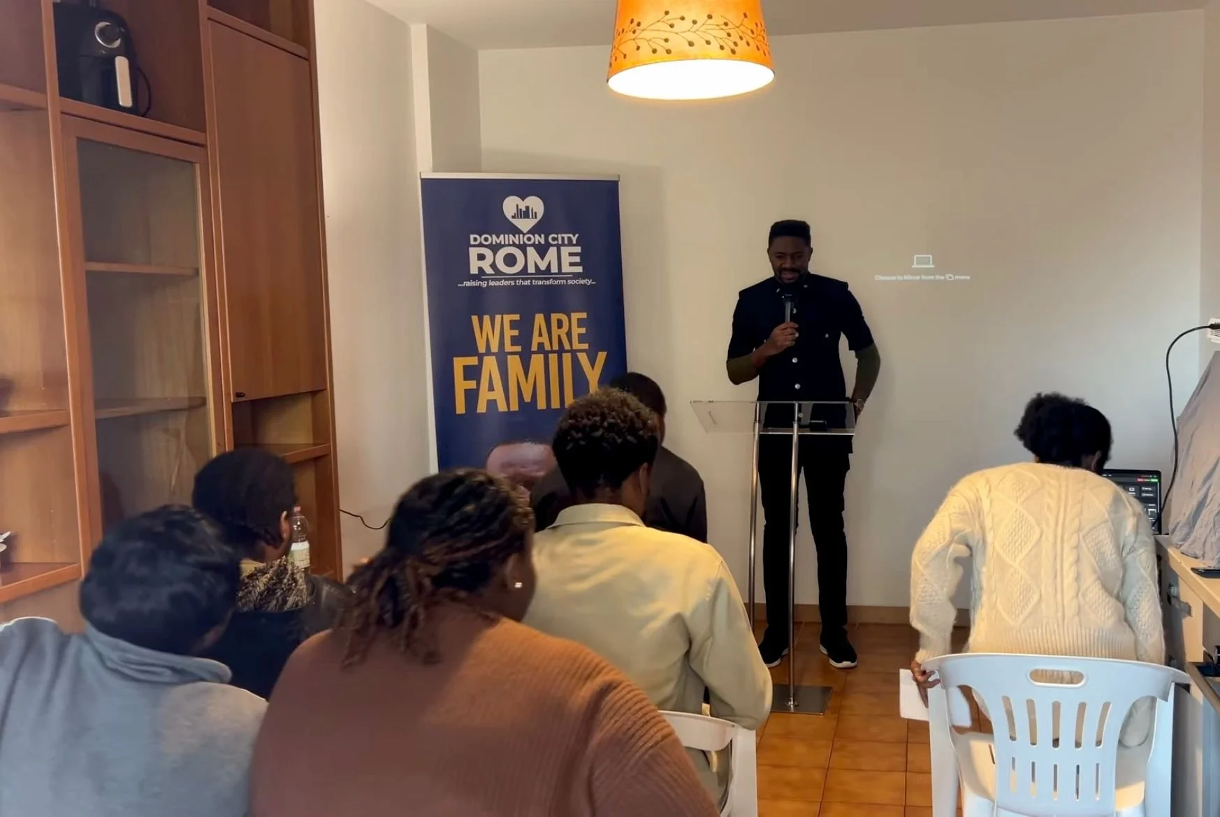 A man is giving a presentation in a room filled with people sitting in chairs, some taking notes or working on laptops. There is a banner that says "DOMINION CITY ROME" and "WE ARE FAMILY". The room has wooden shelves on the left and a ceiling light 