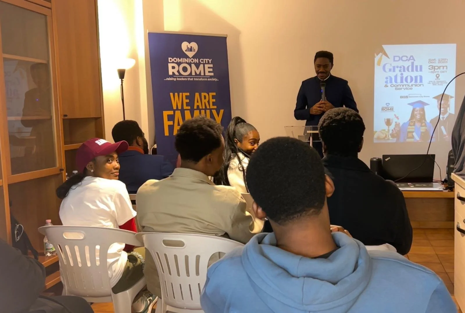 A group of people attending a presentation or seminar in a room. A man stands at the front near a screen, speaking. The screen displays text about a graduation and communion service. A blue banner reads 'DOMINION CITY ROME... WE ARE FAMILY'.