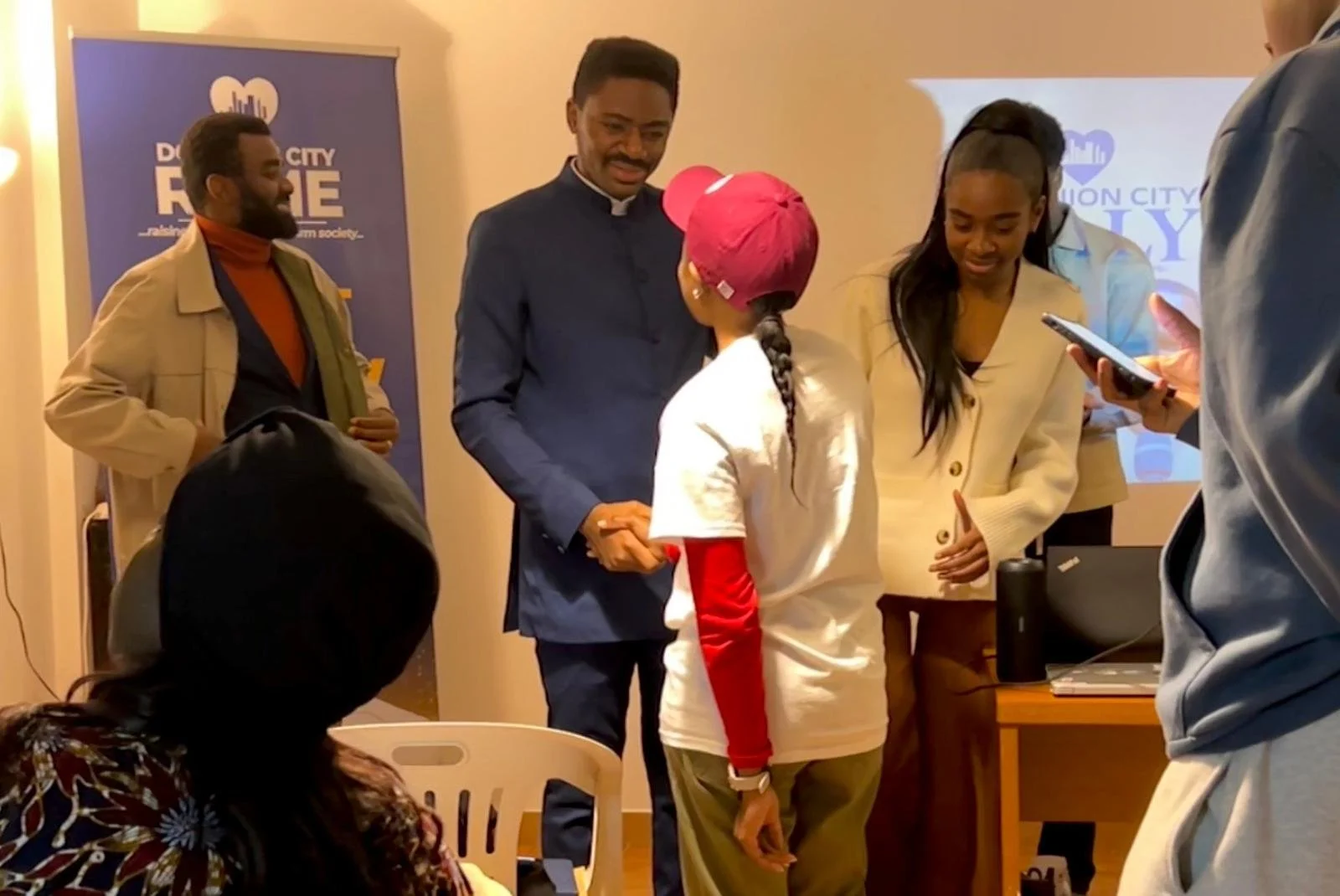 People at an indoor event, including a girl with braided hair and a pink cap shaking hands with a man in a blue suit, while others look on and use their phones.