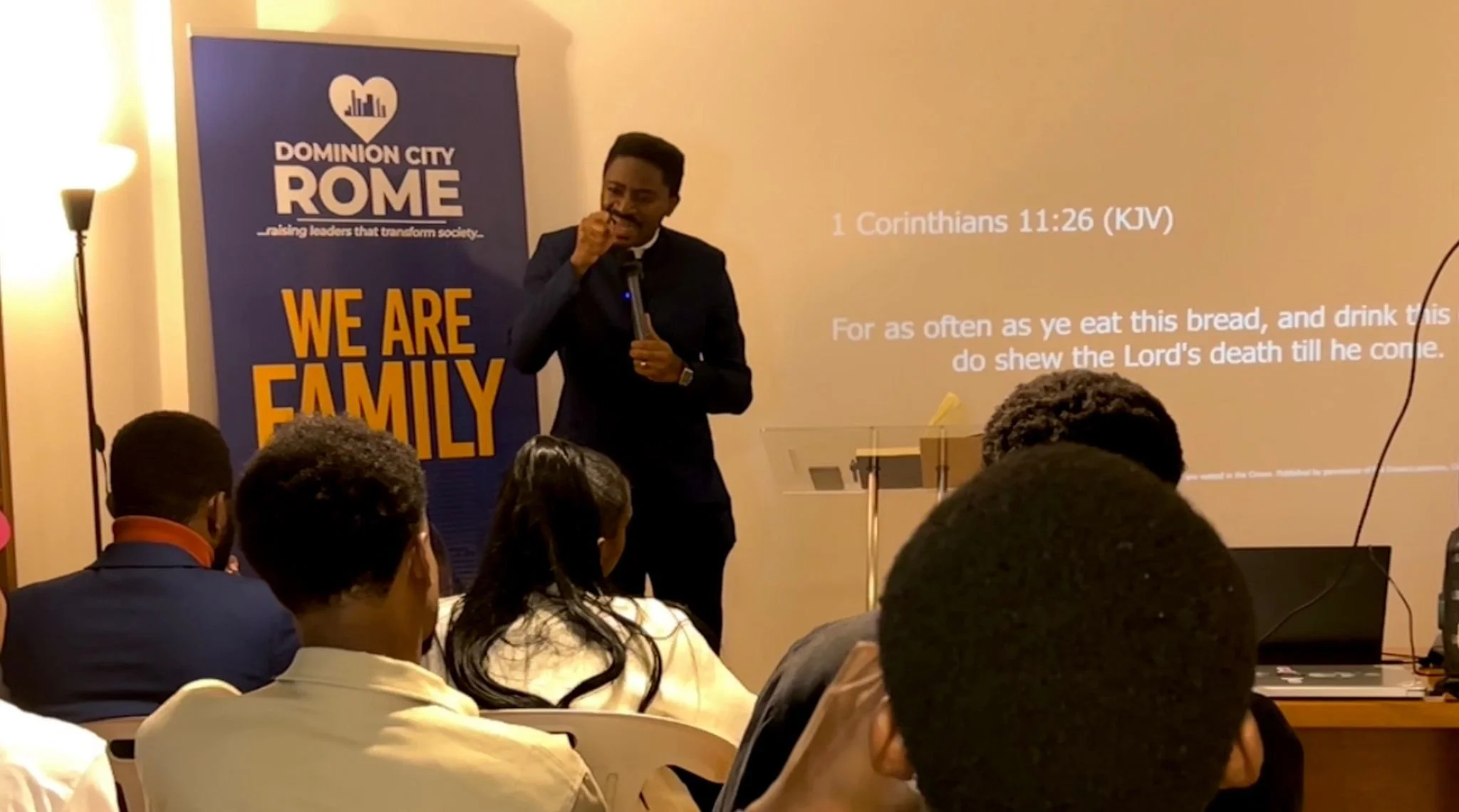 A man in a dark suit speaking into a microphone in front of a screen displaying scripture and a banner that reads 'Dominion City Rome, raising leaders that transform society,' with a mostly adult audience seated and listening.
