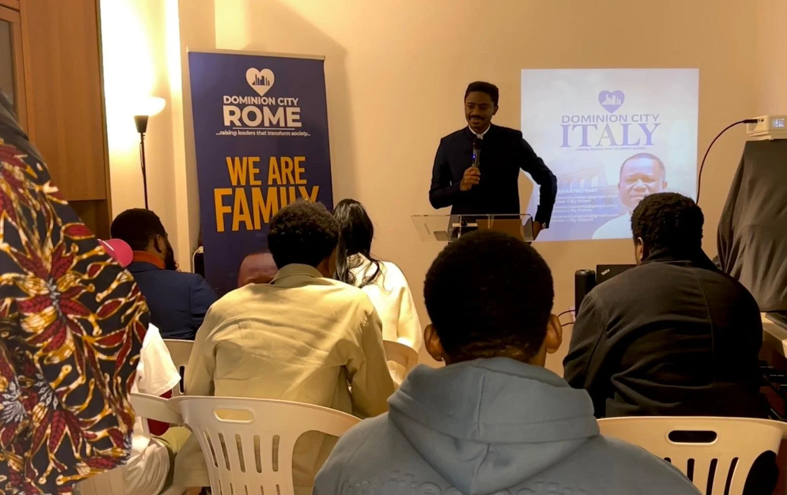 A man in a suit giving a speech at a podium during a community event. Behind him, a screen displays a presentation slide with the title "Dominion City Italy" and an image of a man. To the left, there is a banner with the words "We Are Family" and "Ra