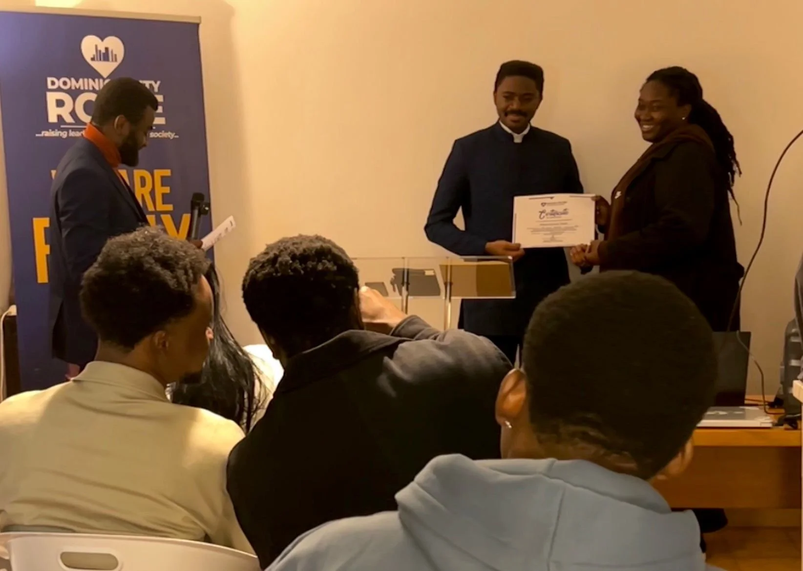A person receiving a certificate from another person during an award ceremony, with several attendees seated and watching. There is a banner in the background that reads "DOMINION CITY RACE" and features a website URL and slogan about making leadersh