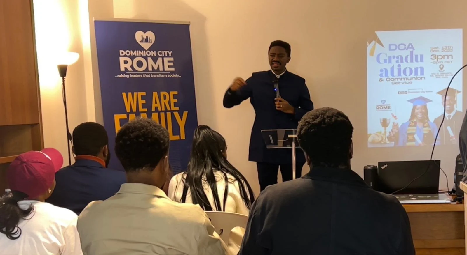 A man wearing a dark jacket giving a speech at a seminar or presentation in a room. Audience members are sitting in front, and a large banner with the words "DOMINION CITY ROME WE ARE FAMILY" is visible. A slide or screen displays information about a