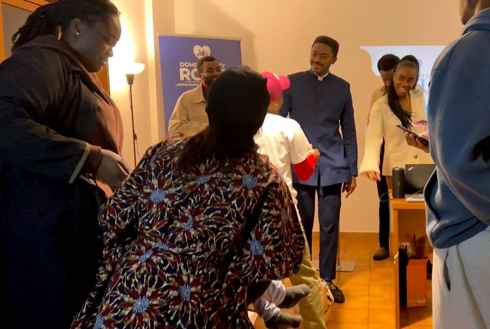 A group of people in a room socializing and smiling, with some looking at their phones. There is a banner in the background with the text 'DOMINION' and 'ROO' visible.