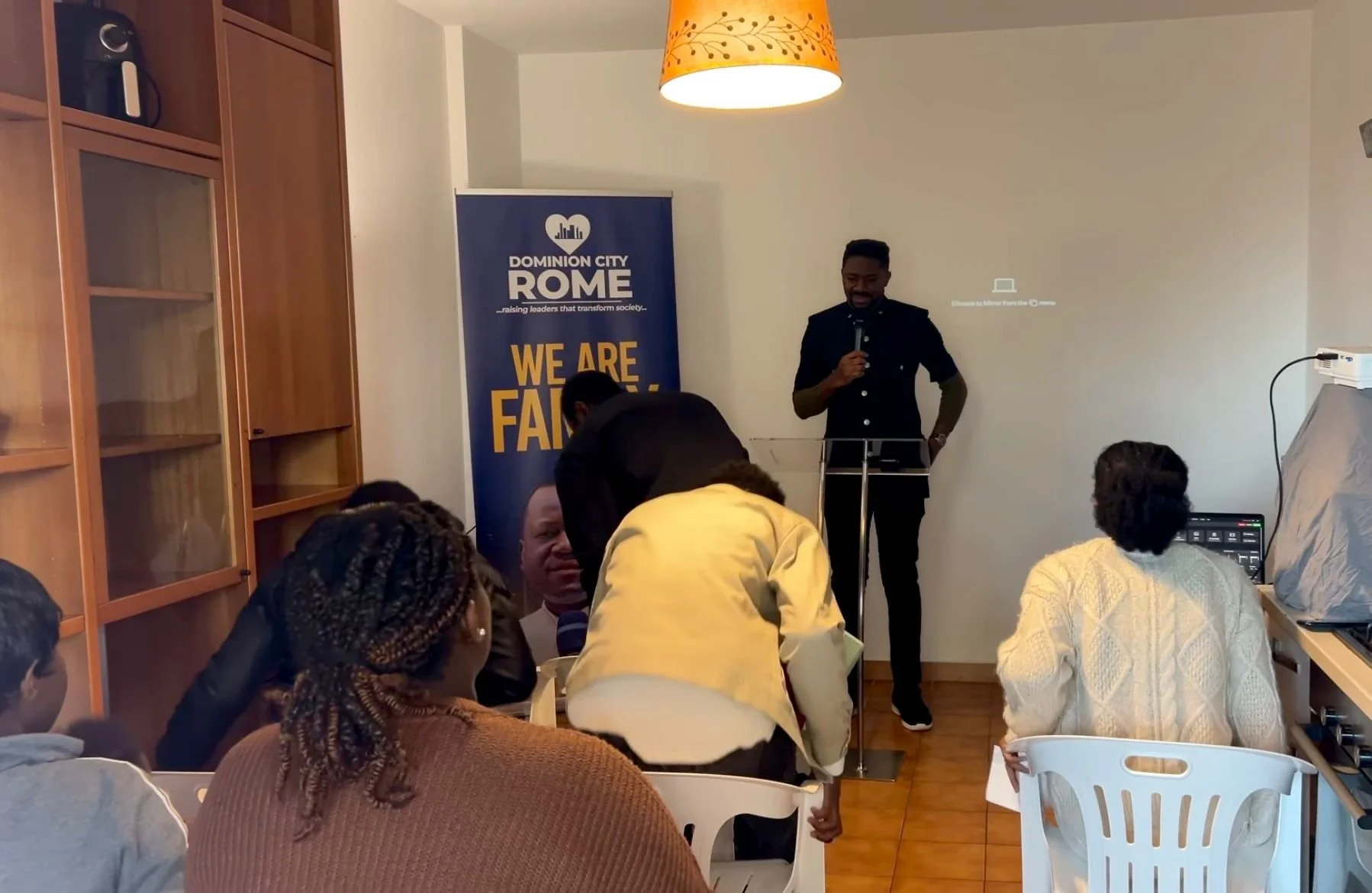 Indoor meeting or workshop with people seated and a speaker standing at a podium. There is a pull-up banner with the words 'DOMINION CITY ROME' and 'WE ARE FAMILIAR', students or participants are sitting in white chairs, some engaging with their lapt