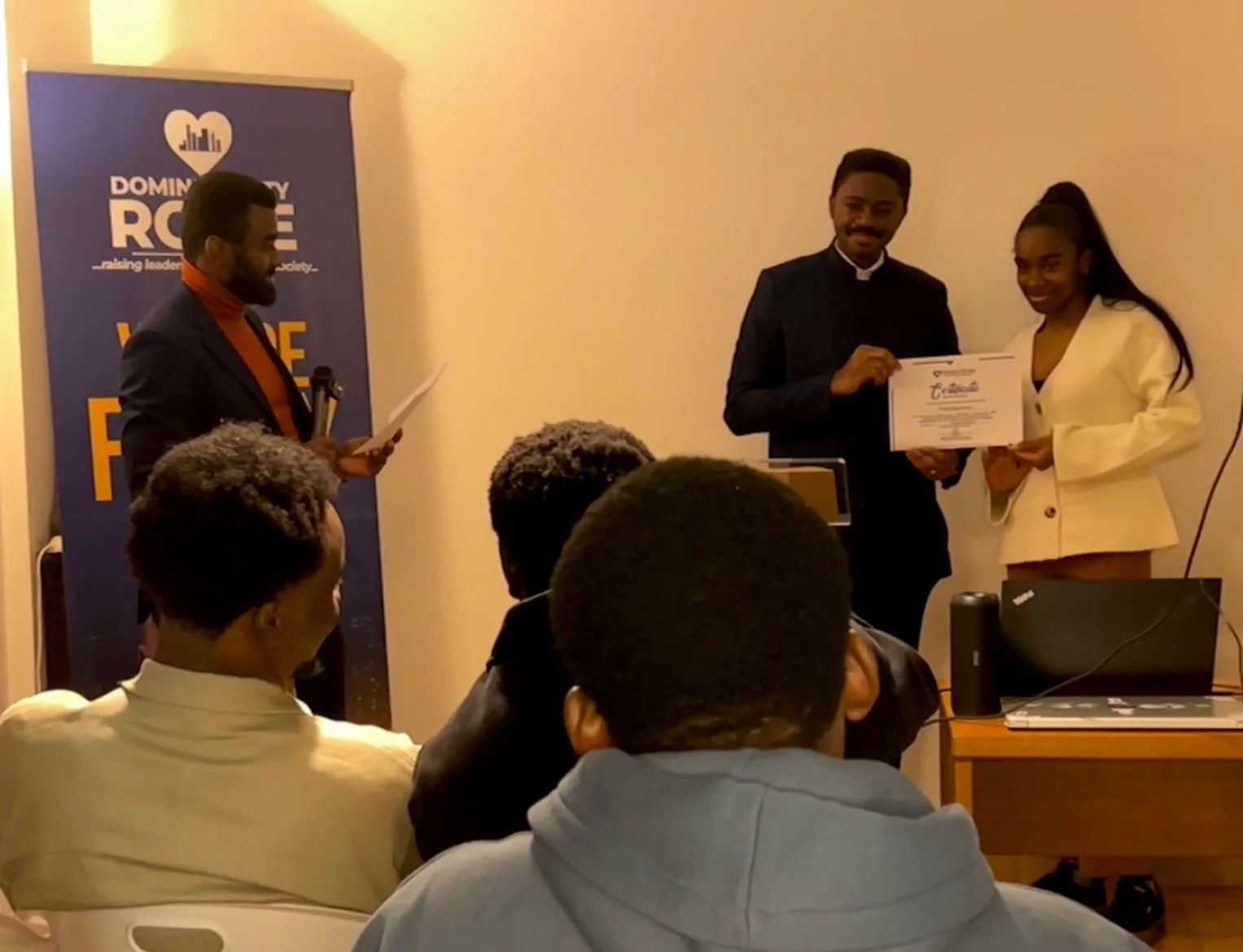 A woman in a white blazer receiving a certificate from a man in a dark suit during an award ceremony, with a presenter reading from a paper and an audience watching.