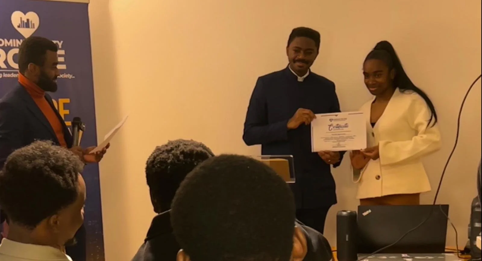 A woman in a cream-colored blazer receiving a certificate from a man in a black suit during an indoor event, with several people seated watching.