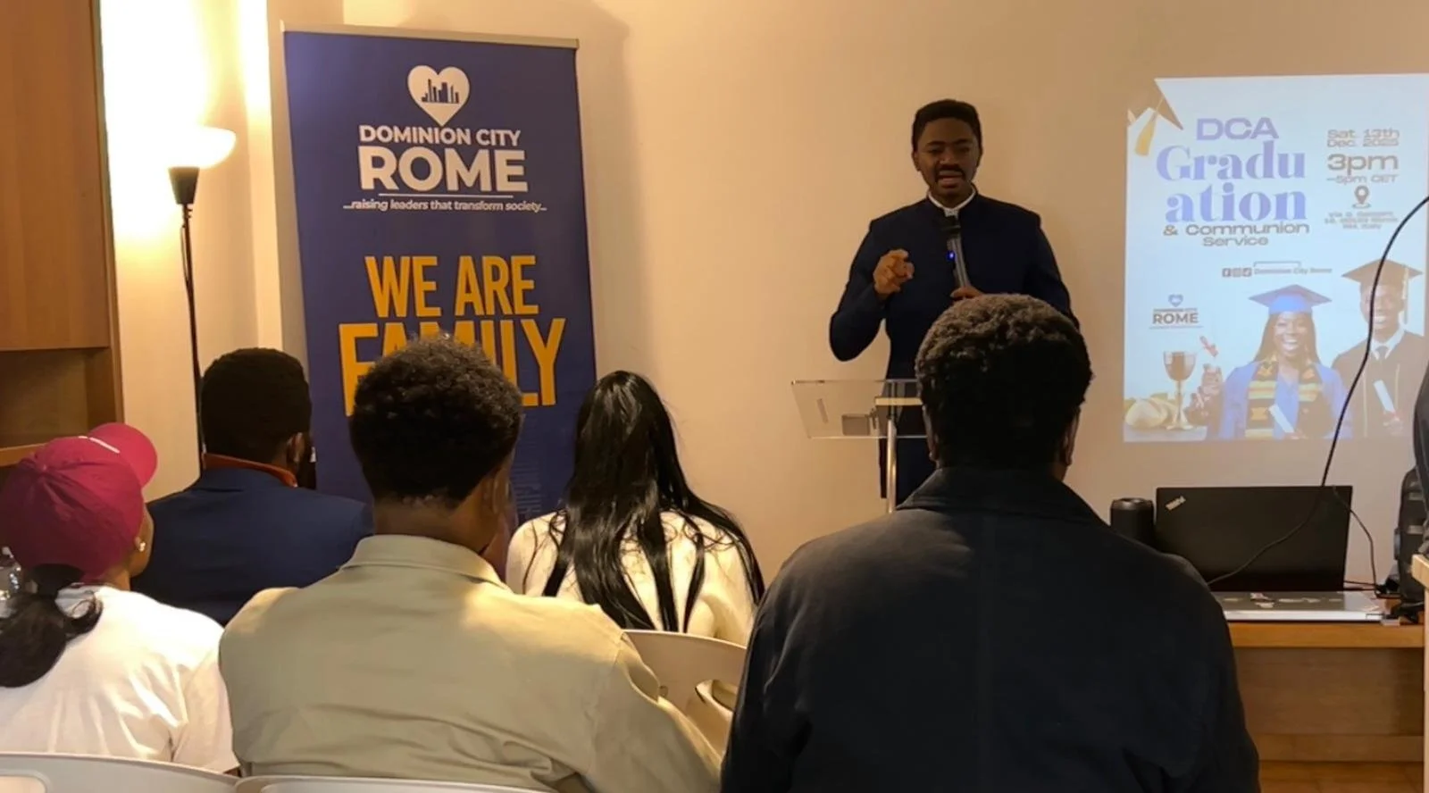 A speaker in a dark suit presenting at an event with an audience seated in front. A large banner with the words 'We are family' and 'Dominion City Rome' is behind the speaker, along with a projected slide showing graduation and communion service info