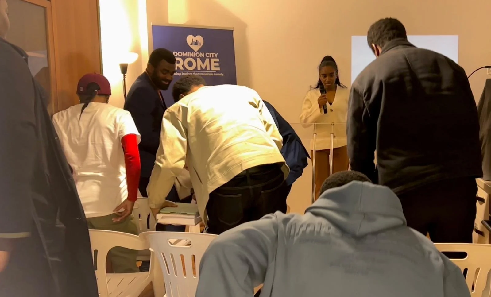 A group of people gathered around a table in a meeting room, with a woman standing at a podium holding a microphone. There is a banner in the background that reads "Dominion City Rome."
