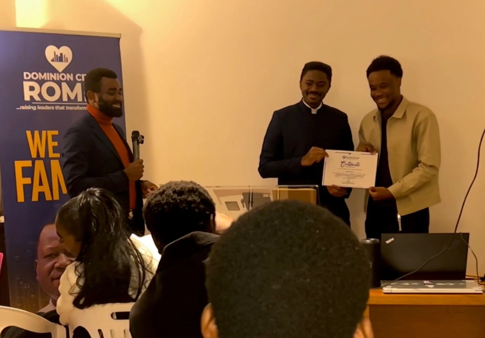 Three men standing in front of an audience at an indoor event. One man is holding a certificate and smiling, while another man is presenting it. The third man is speaking into a microphone. A banner with the logo and text 'DOMINION CITY ROMA' and 'We