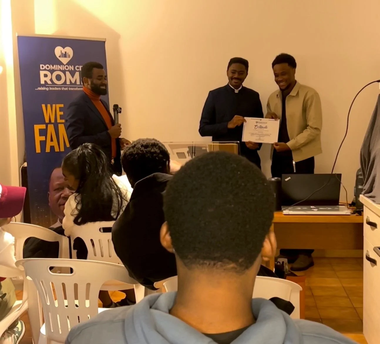 A man in a dark blazer presents a certificate to a young man in a beige jacket at an indoor event, with an audience and a banner in the background.