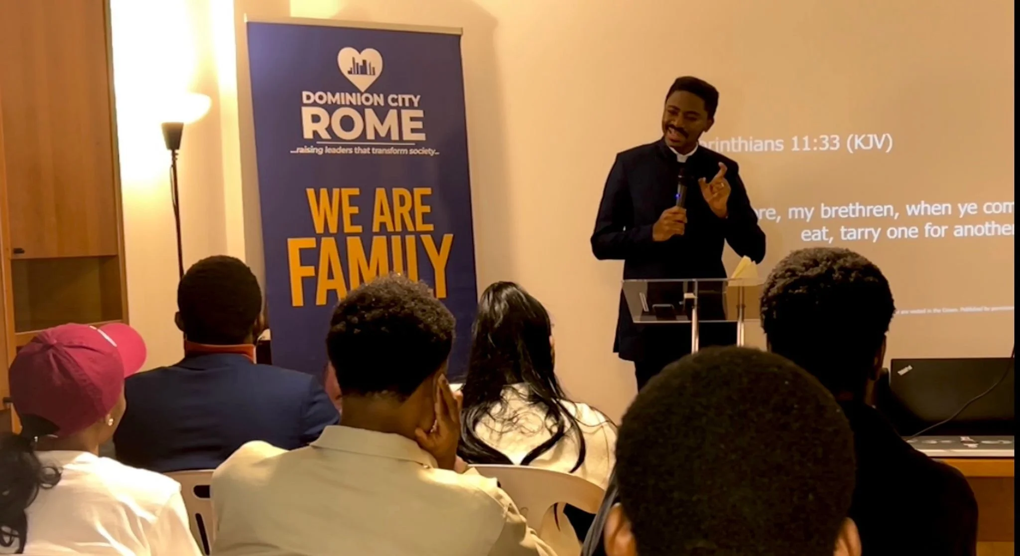 A man dressed in black standing behind a podium, speaking to an audience. Behind him is a blue banner that reads 'Dominion City ROME' with a tagline underneath. The audience is seated, facing the speaker, with some taking notes or listening attentive
