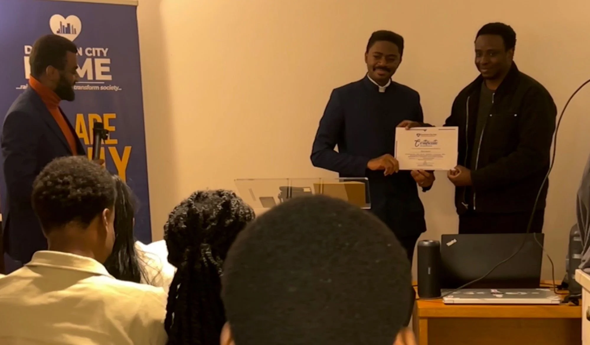 Three men standing indoors, with one man presenting a certificate to another, while a woman and another man watch, in front of a blue banner with white text and logo.