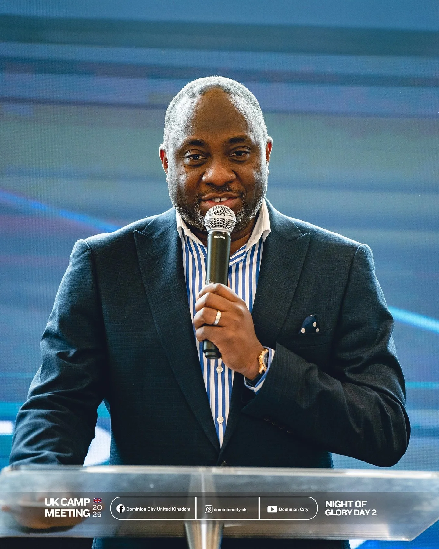 A man in a dark suit holding a microphone and speaking at a conference or event with a blue background. Text overlay indicates it's a UK camp meeting in 2025, Night of Glory Day 2, organized by Dominion City, with social media handles for Dominion City United Kingdom.