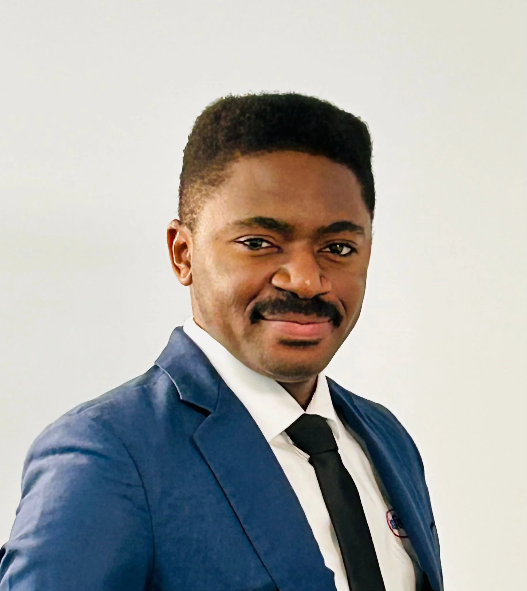 Close-up of a man with a short haircut and mustache, wearing a blue suit, white dress shirt, and black tie, smiling against a plain white wall.