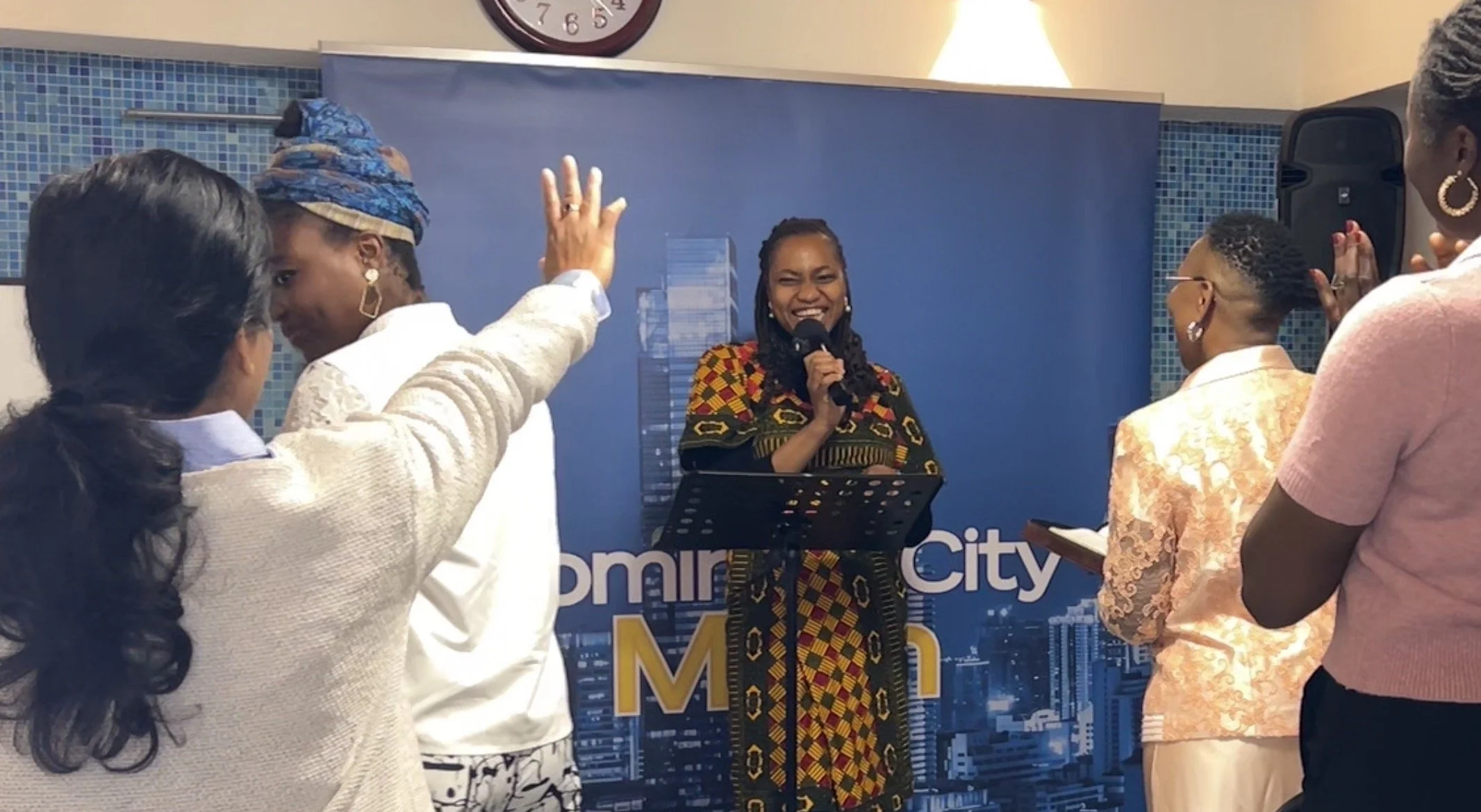 A woman in traditional colorful African attire speaking into a microphone during an event, surrounded by several women in white, pink, and patterned clothing who are listening and participating.