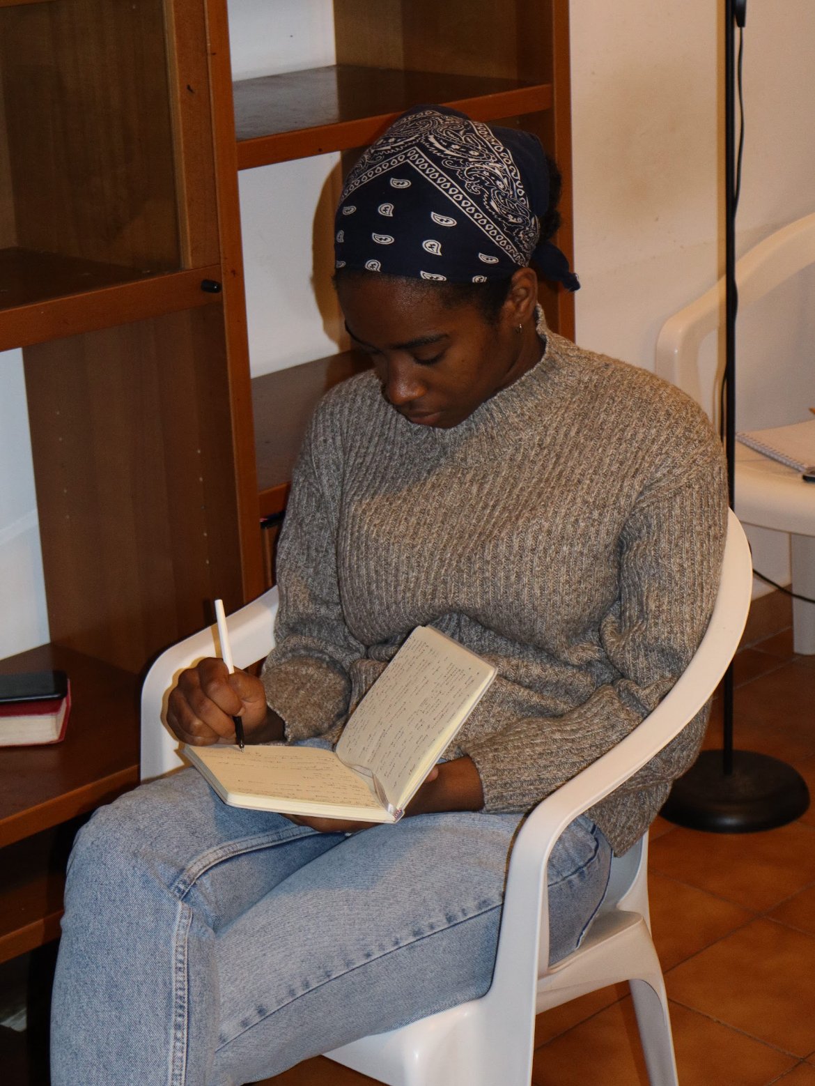 A person sitting in a white plastic chair, writing in a notebook with a pen, wearing a gray sweater and a patterned bandana on their head, near wooden shelves and a side table with books and a notebook.