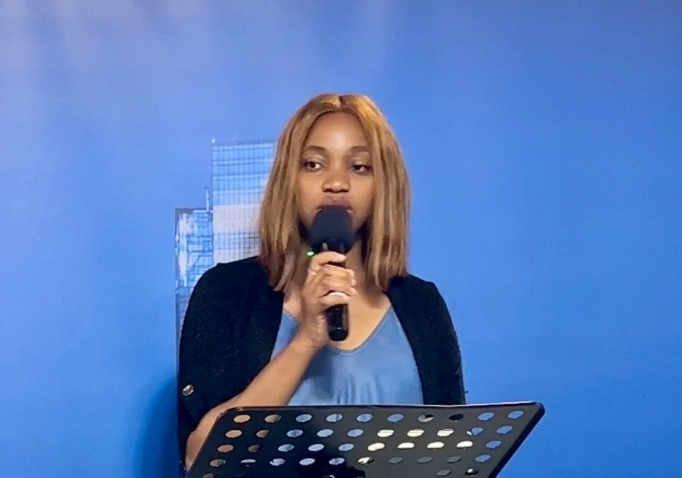 Woman with shoulder-length light brown hair speaking into a microphone at a presentation or event, standing in front of a blue background with a cityscape graphic.