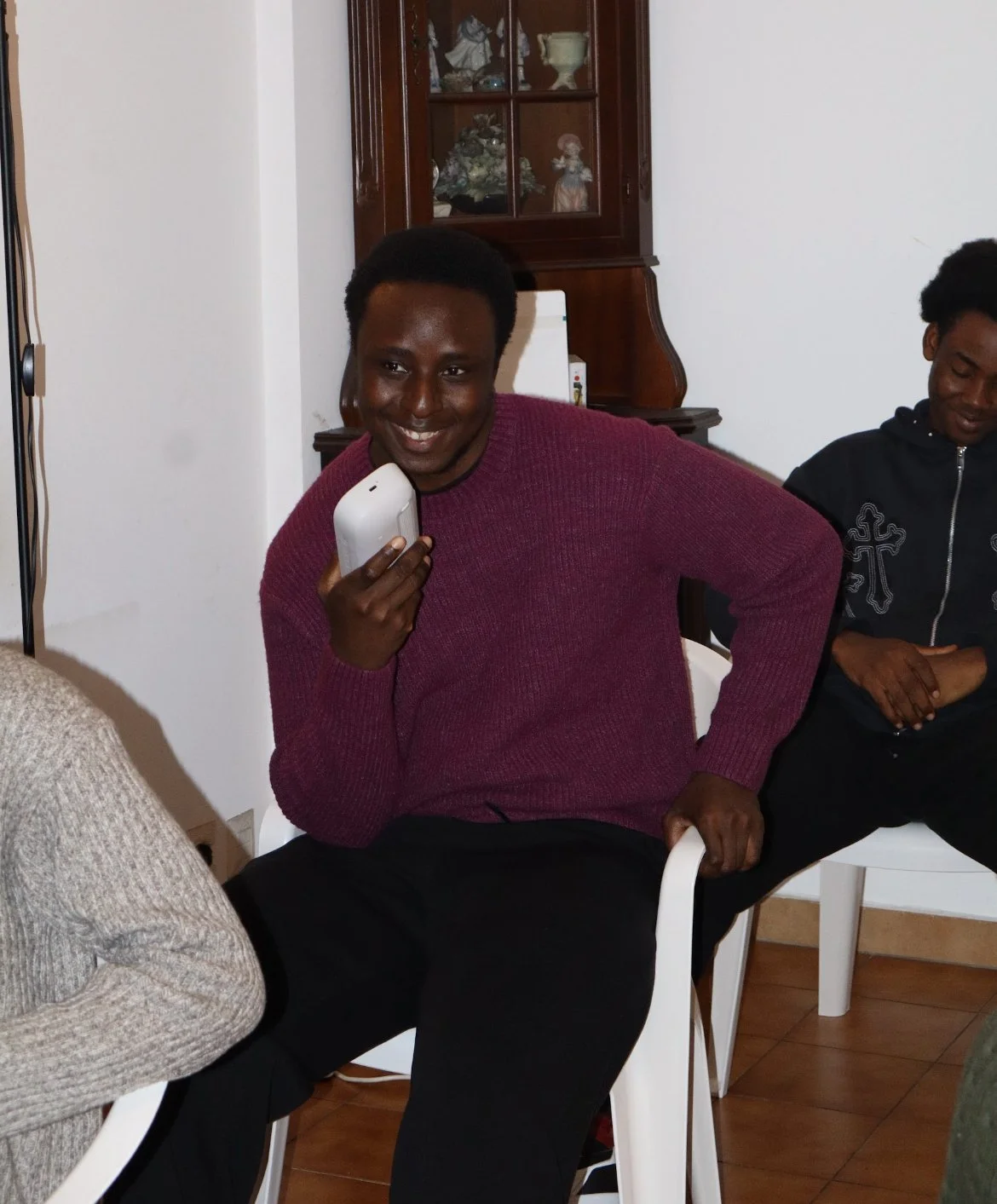 A man in a red sweater smiling while holding a white phone against his cheek, sitting inside a room with wooden furniture and other people.