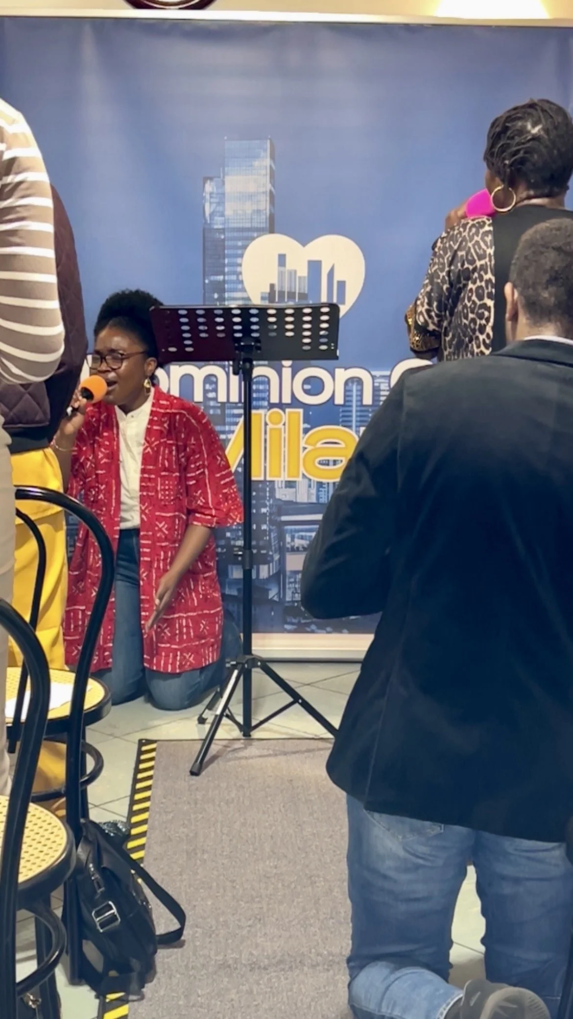A woman wearing glasses and a red patterned shawl is singing into a microphone, kneeling on the floor. There are several people around, and a backdrop with cityscape graphics and the words 'amazing' and 'glia' is in the background.