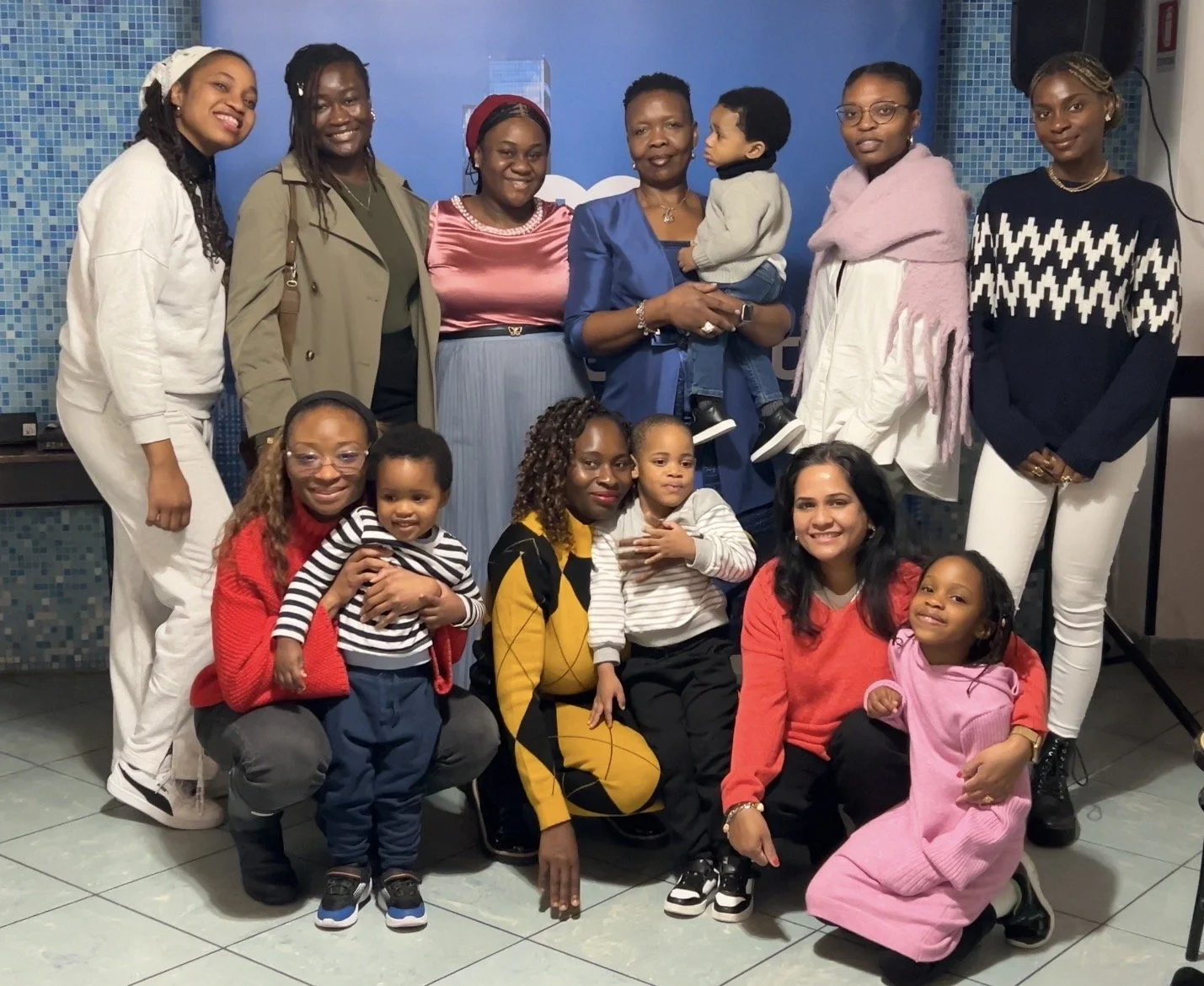 Group of women and children posing together indoors, some sitting and some standing, in front of a blue tiled wall.
