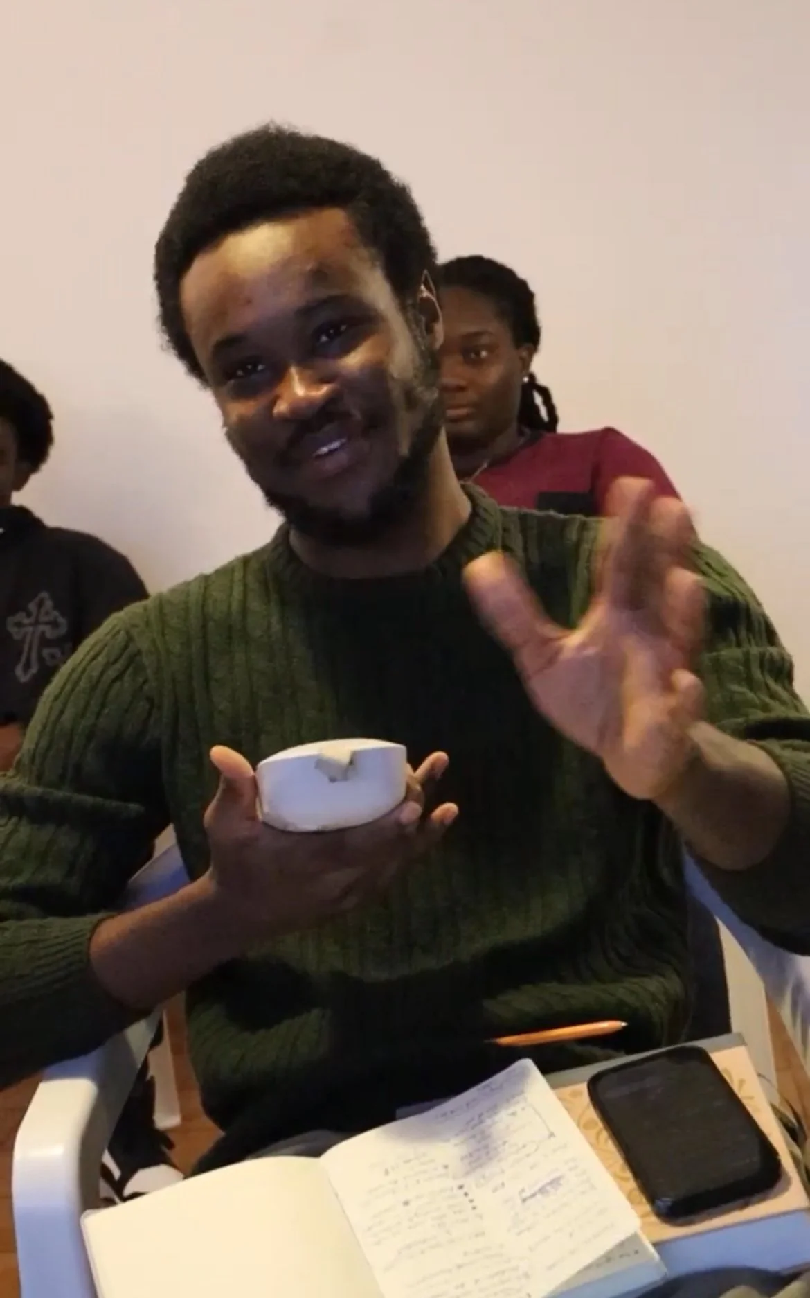 A young man with short curly hair and a beard is smiling and gesturing with his hand, holding a small white bowl with what appears to be a piece of paper or napkin inside. He is seated at a table with an open notebook and a smartphone in front of him