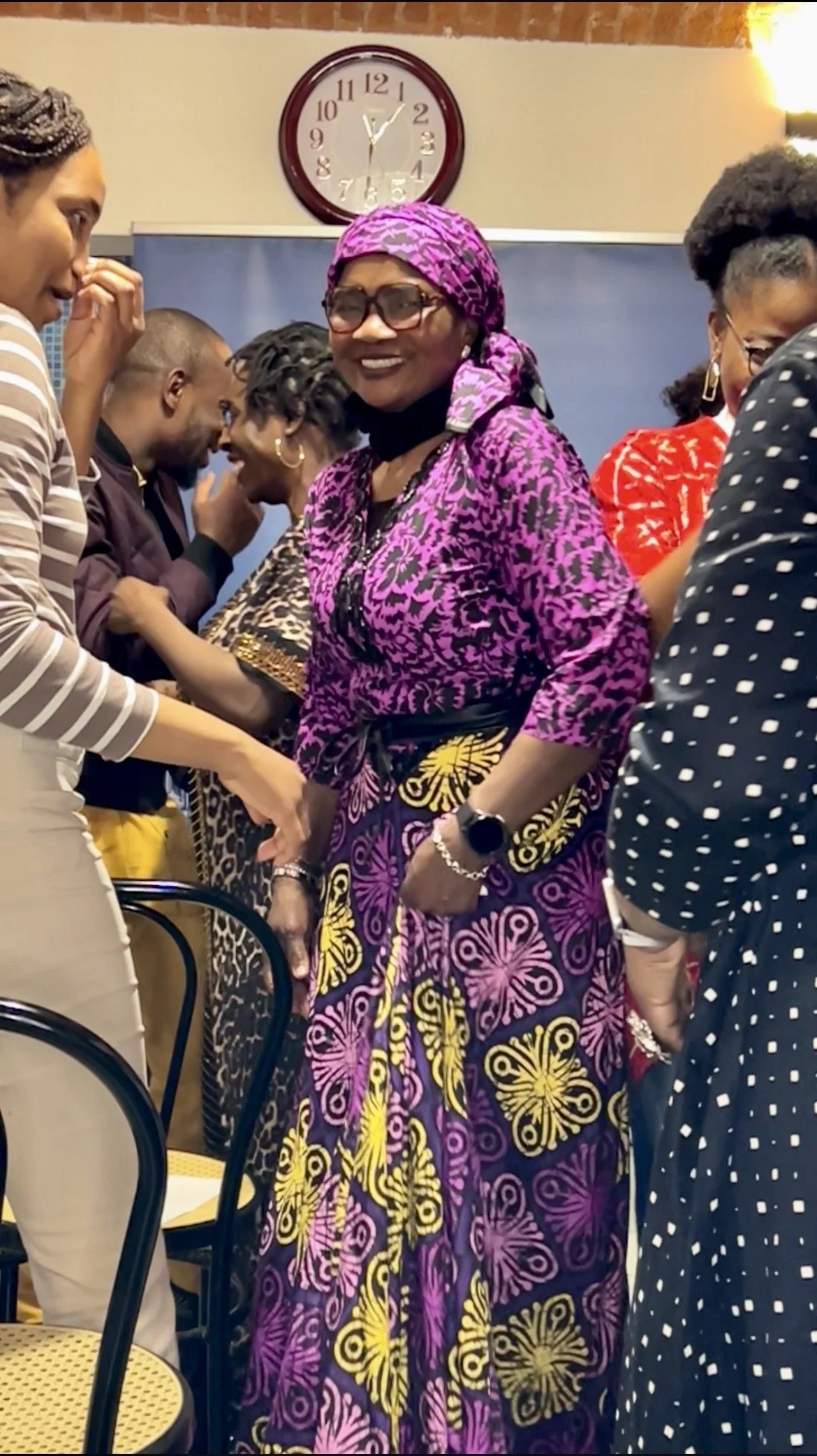 A group of diverse people socializing at a gathering indoors, with a woman in a vibrant purple and yellow butterfly-patterned dress and glasses at the center.