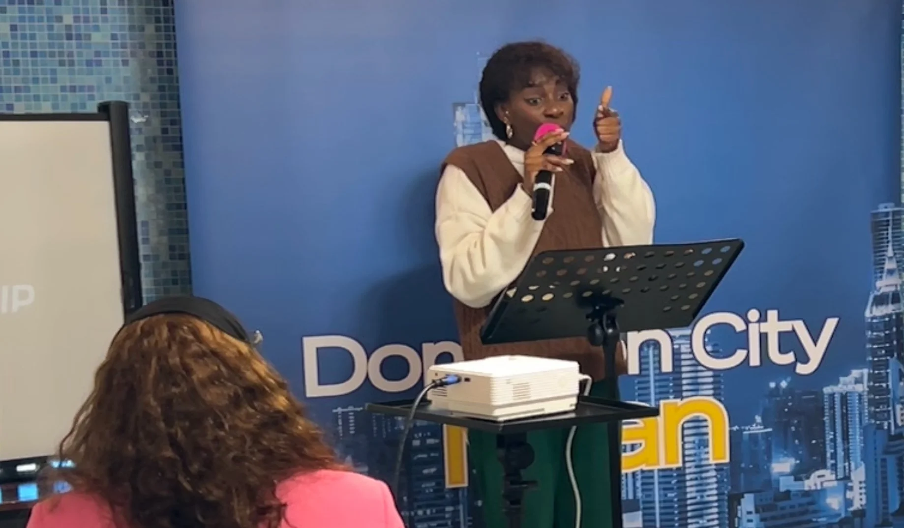 A woman standing at a podium with a blue cityscape background, speaking into a microphone, and giving a thumbs-up with her right hand. There are people seated in front of her, one wearing a pink top and another with curly hair.