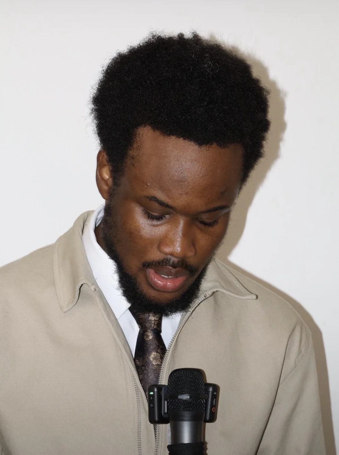 A man with curly black hair and a beard, wearing a beige jacket and patterned tie, is looking down while speaking into a microphone.