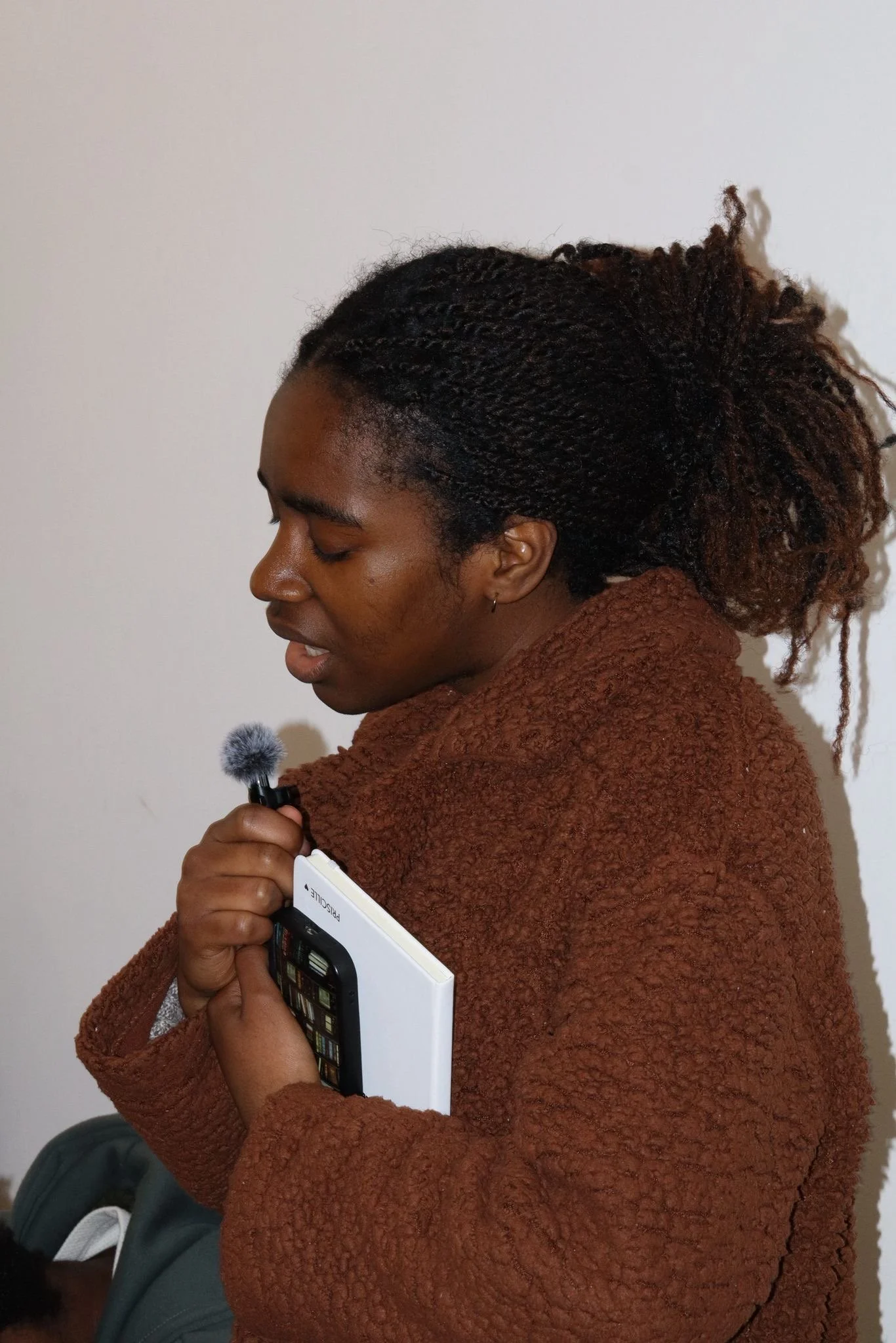 A woman with dark curly hair tied in a loose ponytail, wearing a brown textured hoodie, is holding a small microphone and a notebook, standing against a plain white wall.