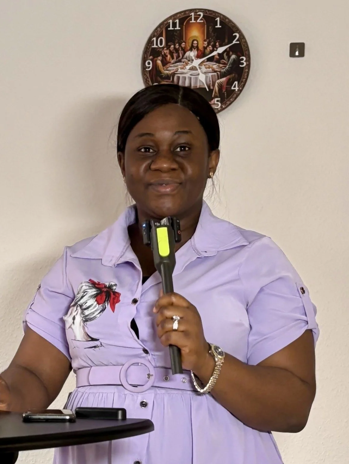 A woman standing in front of a wall clock with a painting of 'The Last Supper' and holding a microphone, wearing a purple dress with a cartoon chef figure on the chest.