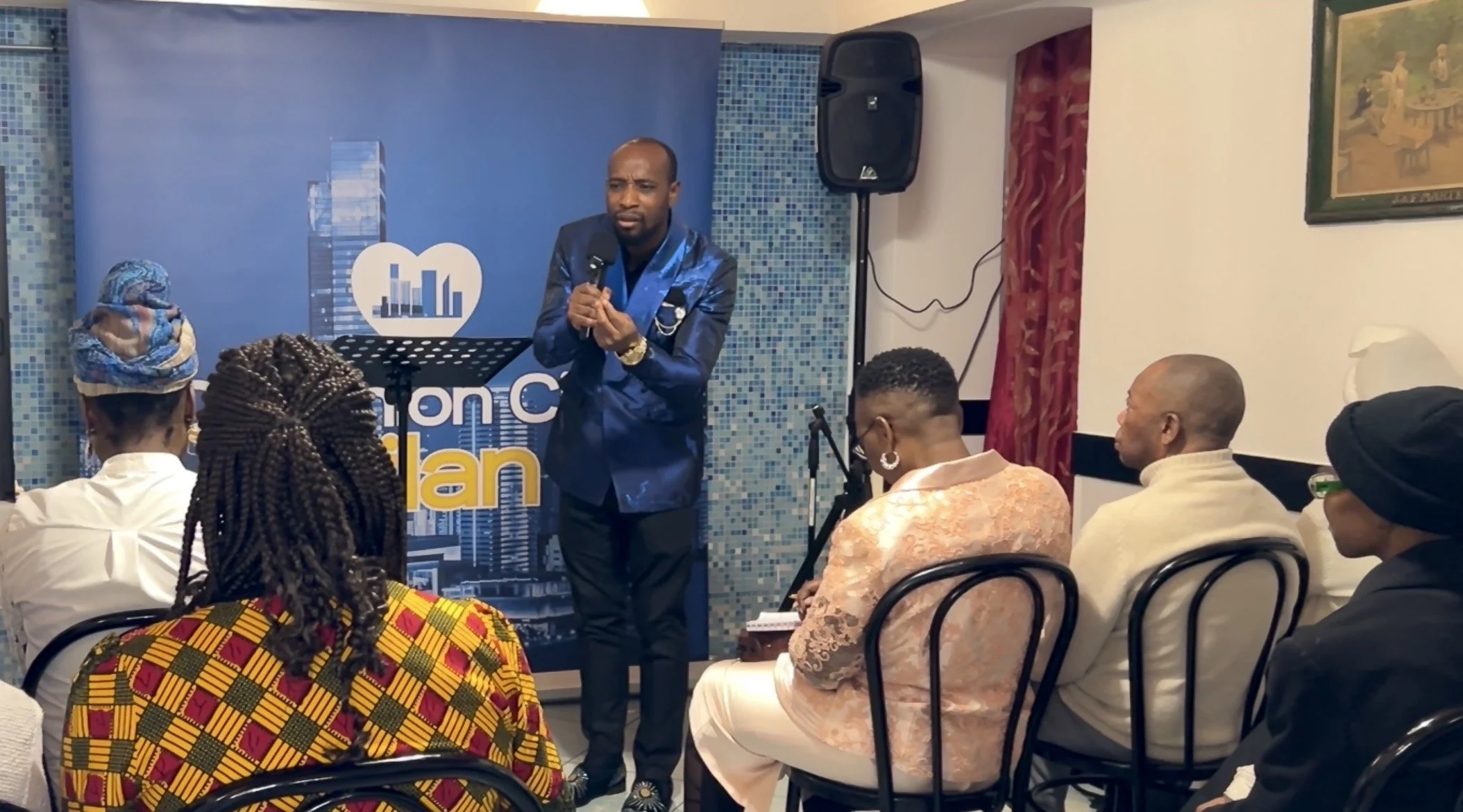 A man in a blue suit speaking into a microphone at an indoor event with seated attendees. Behind him is a blue backdrop with a cityscape and heart logo, and a sign that reads 'Lions Club of ...'. Audience members, mostly women, are listening attentiv