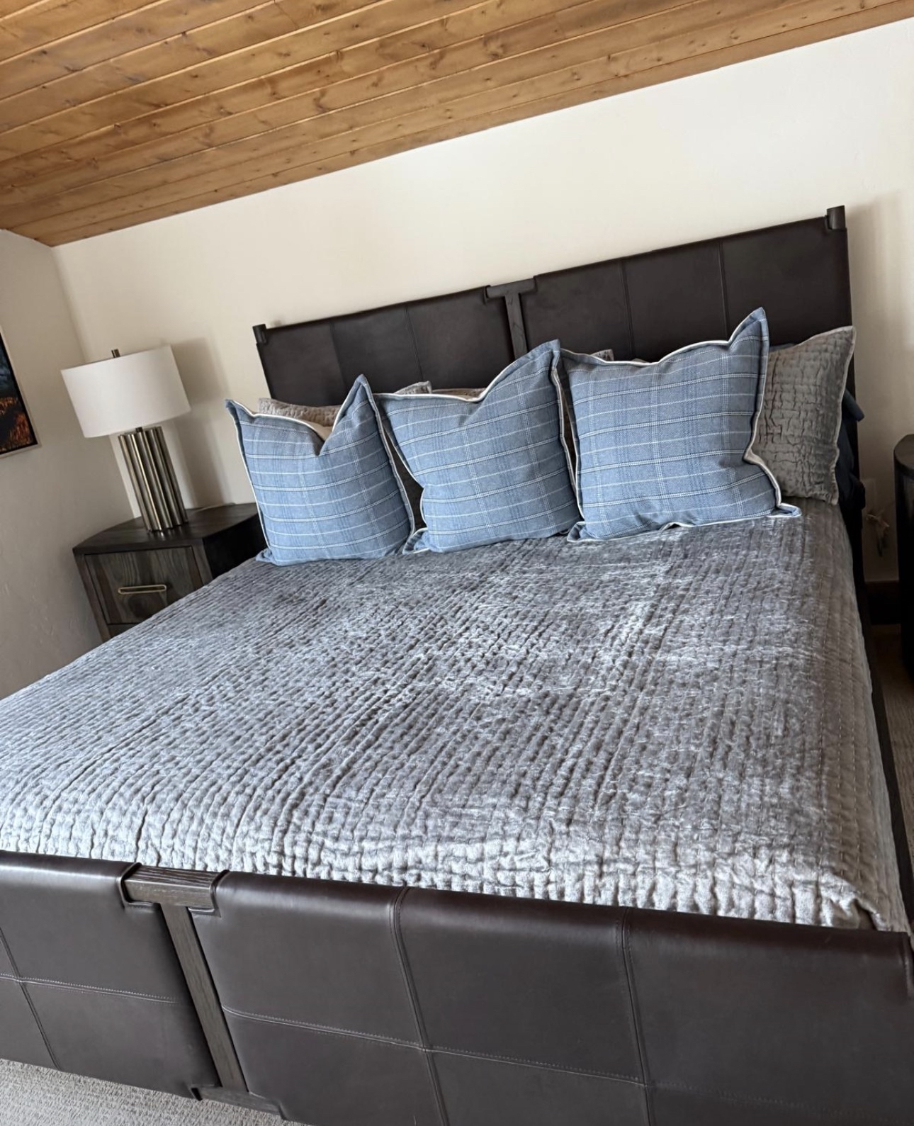 A neatly made bed with a gray velvet quilt and four blue plaid pillows, topped with a gray textured pillow, in a bedroom with a wooden ceiling and a nightstand with a lamp.