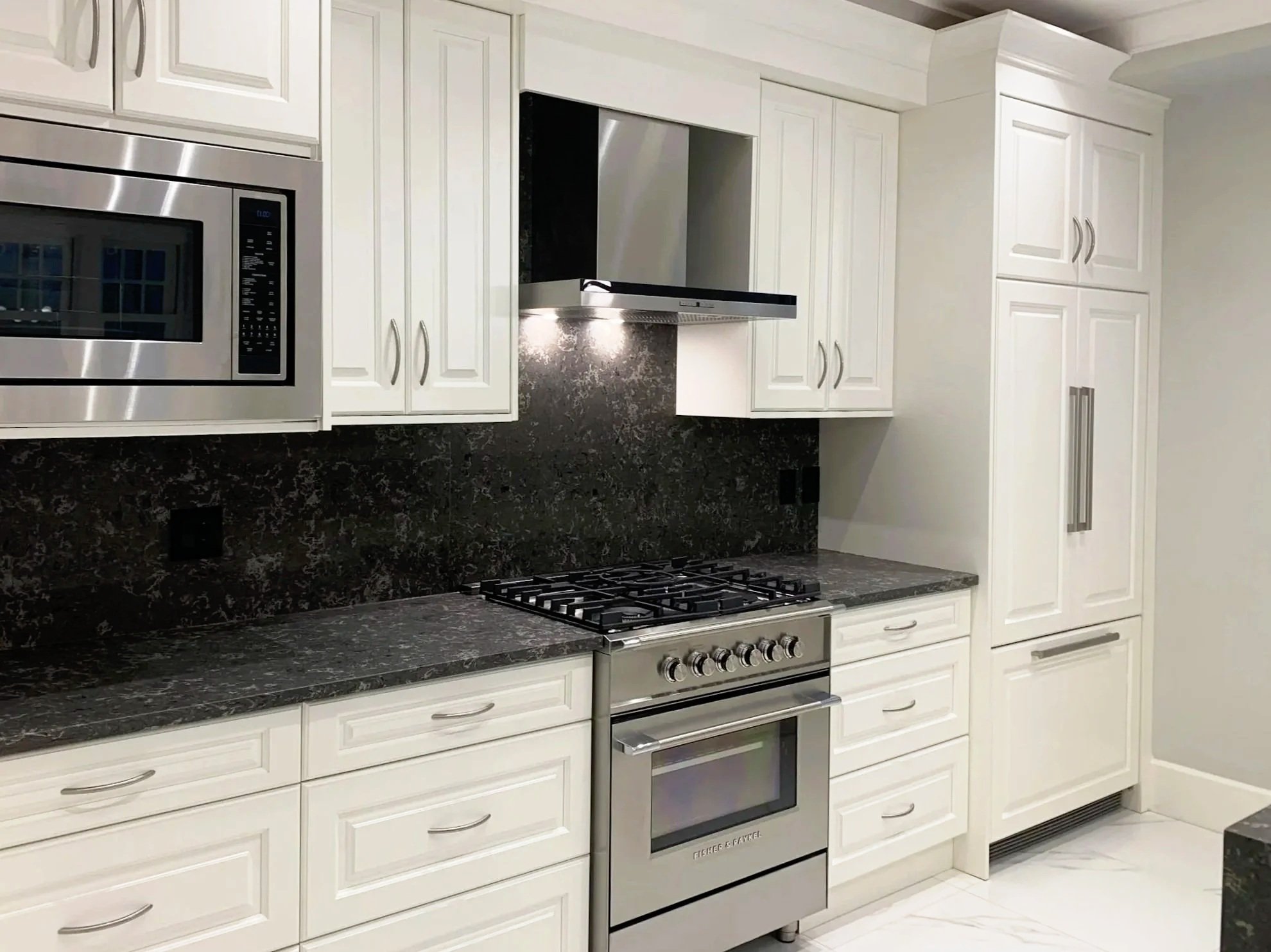 White kitchen cabinets with silver handles, black and gray granite countertops, stainless steel microwave, stove with oven, and range hood. The floor is white with gray marble veining.
