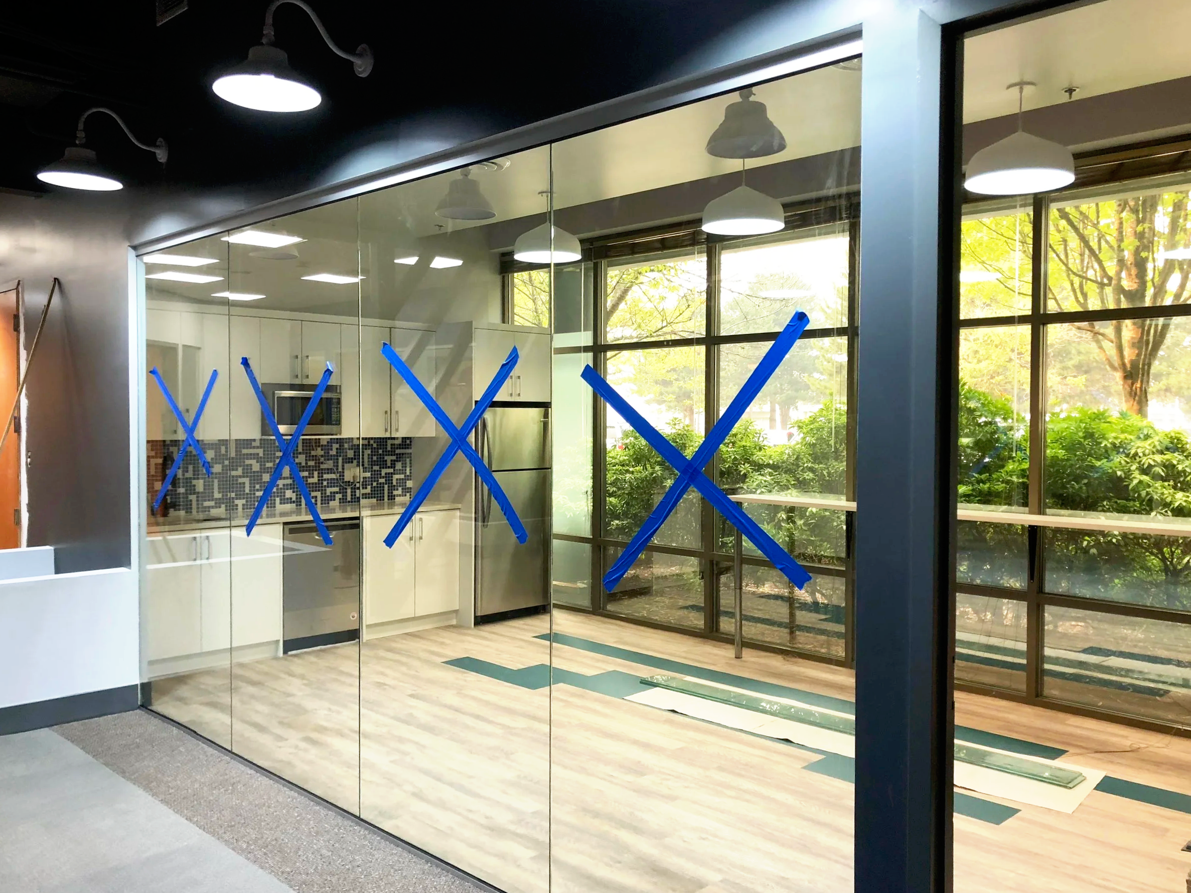 Interior of an office or commercial space with large glass windows, white ceiling with modern hanging light fixtures, and a partial view of a kitchenette or workspace area with white cabinets and geometric backsplash. Blue painter's tape forms X shapes on the glass, indicating ongoing renovations or cleaning.