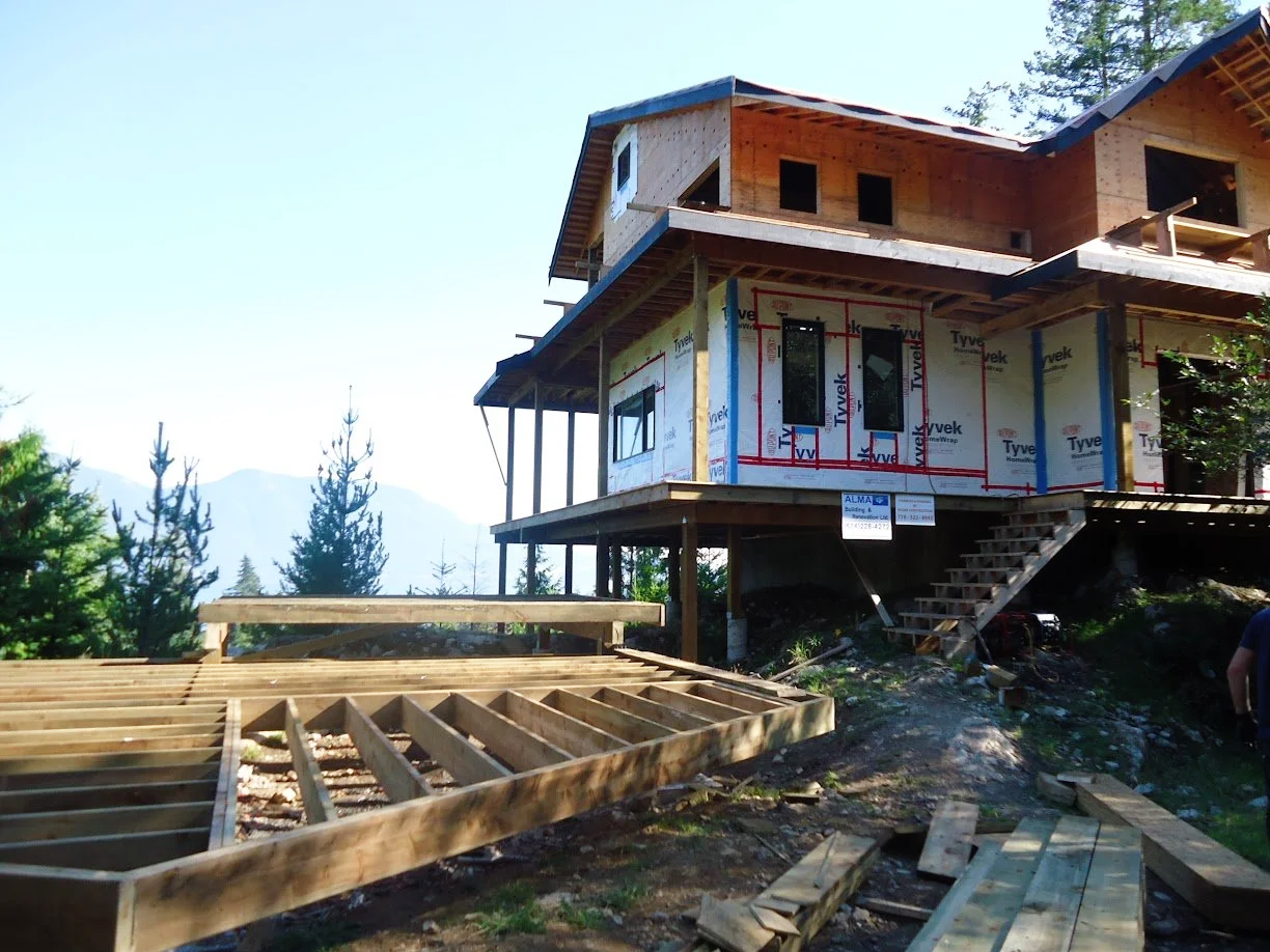 Large house under construction with wooden framing and a partially built deck, surrounded by trees and mountains in the background.