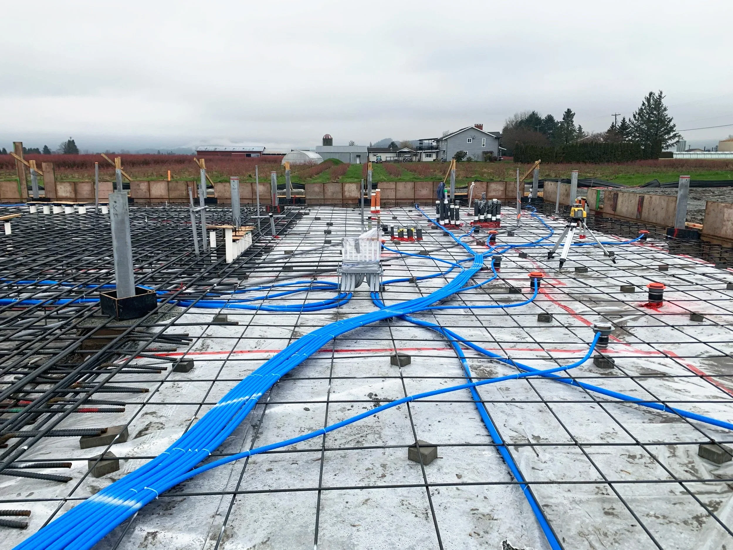 Construction site with exposed rebar and blue plumbing pipes, with a rural landscape in the background including houses, fields, and trees under overcast skies.
