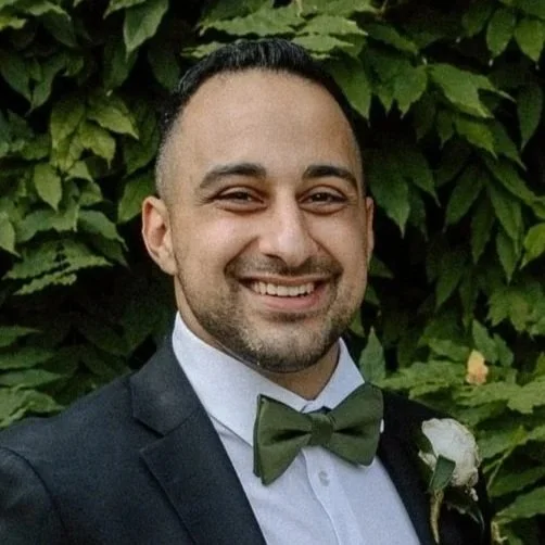 Chris Chebaya smiling in front of green leafy background, wearing a tuxedo with a white shirt, green bowtie, and boutonniere.