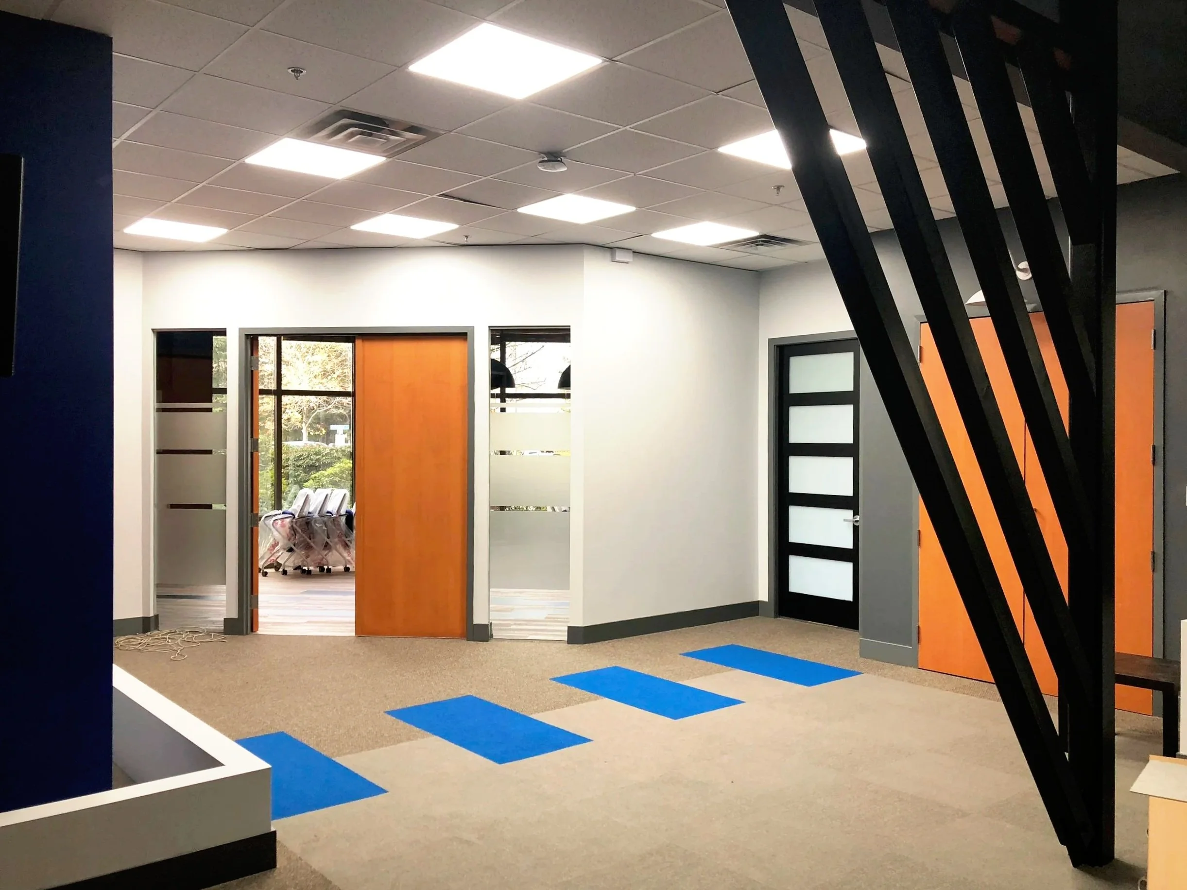 Interior of an office space with open door, black and orange wall decor, tiled ceiling with bright fluorescent lights, beige carpet with blue geometric shapes, and a view into a room with windows and chairs.