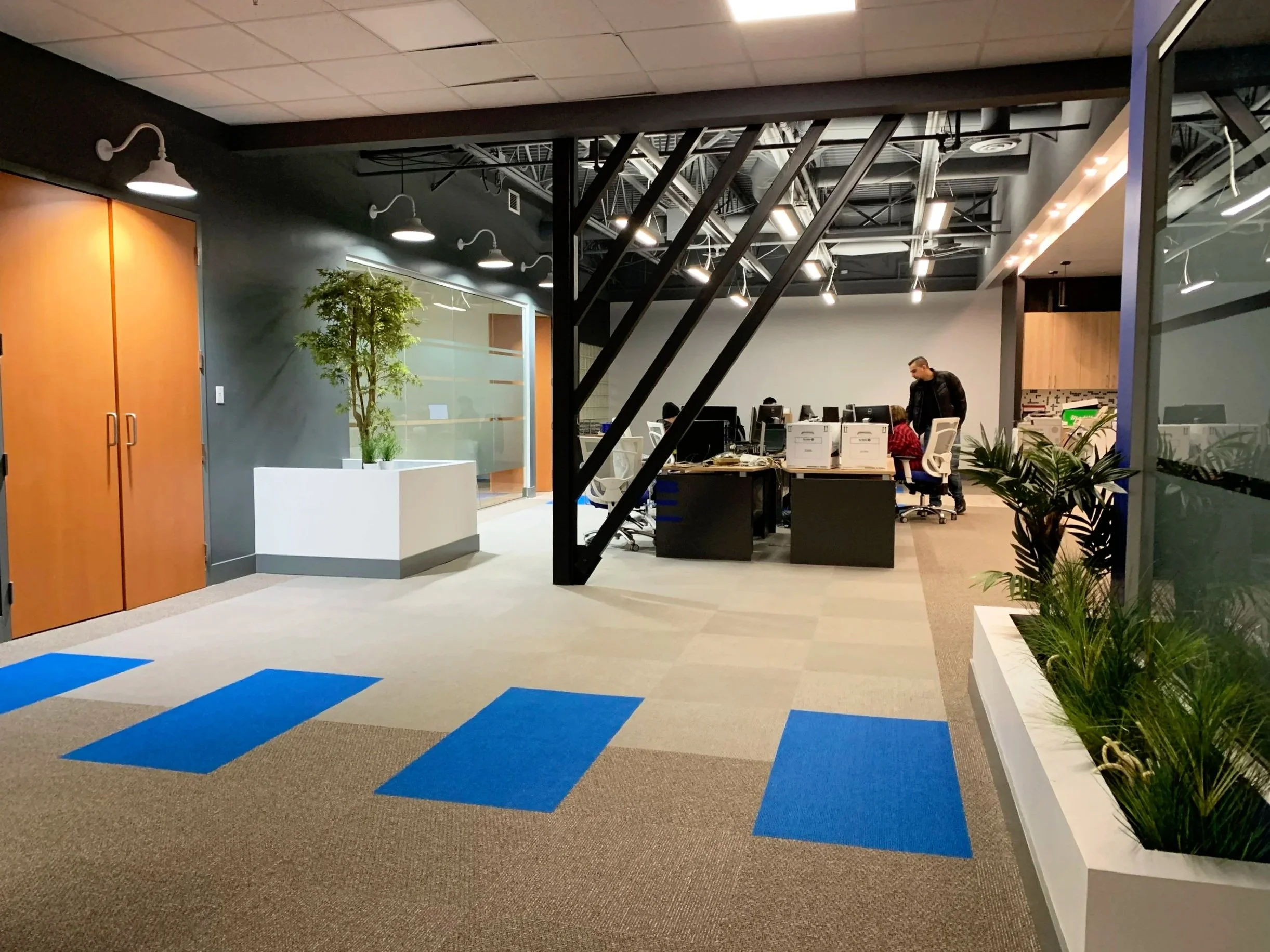 An office interior with blue carpet tiles, plants, wooden doors, and desks with computers. People are working at desks while a man is walking past them.