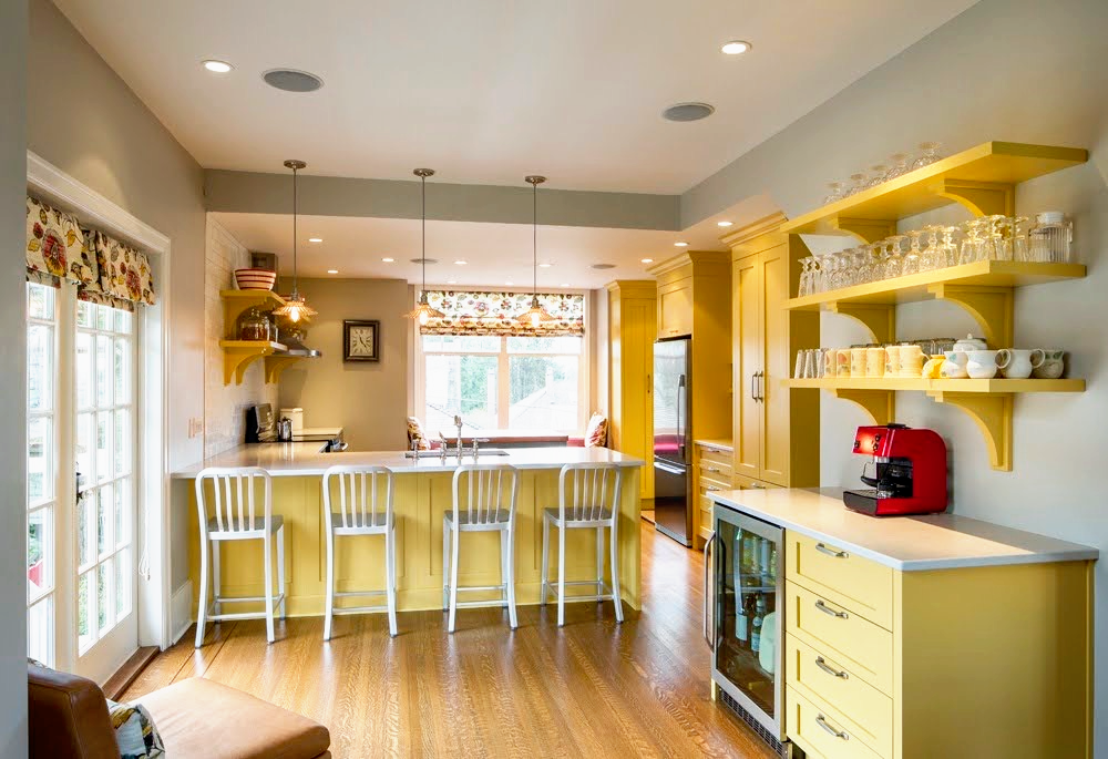 Bright kitchen with yellow cabinets, open shelves holding glassware and dishes, a red coffee machine on the counter, and a breakfast bar with four white chairs. Large window provides natural light.