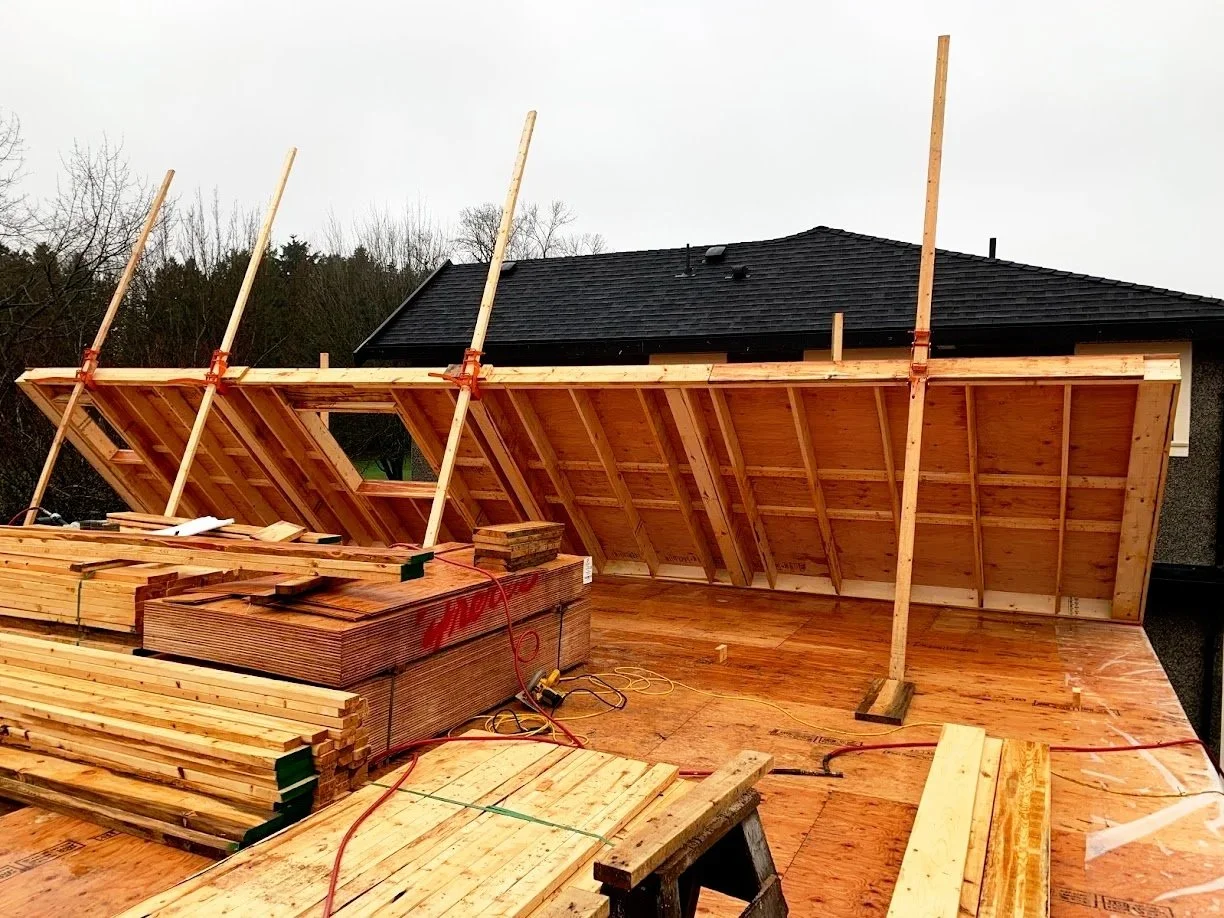 Construction site on a residential roof with new wooden framework being built, including plywood and lumber stacked nearby.