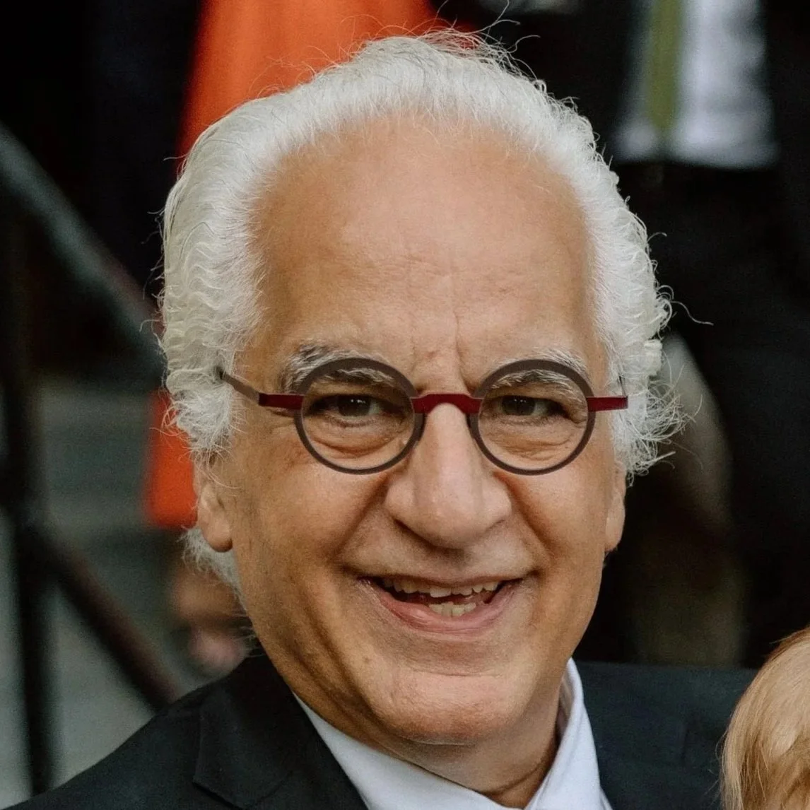 Tony Chebaya, wearing round glasses with black and red frames, smiling.