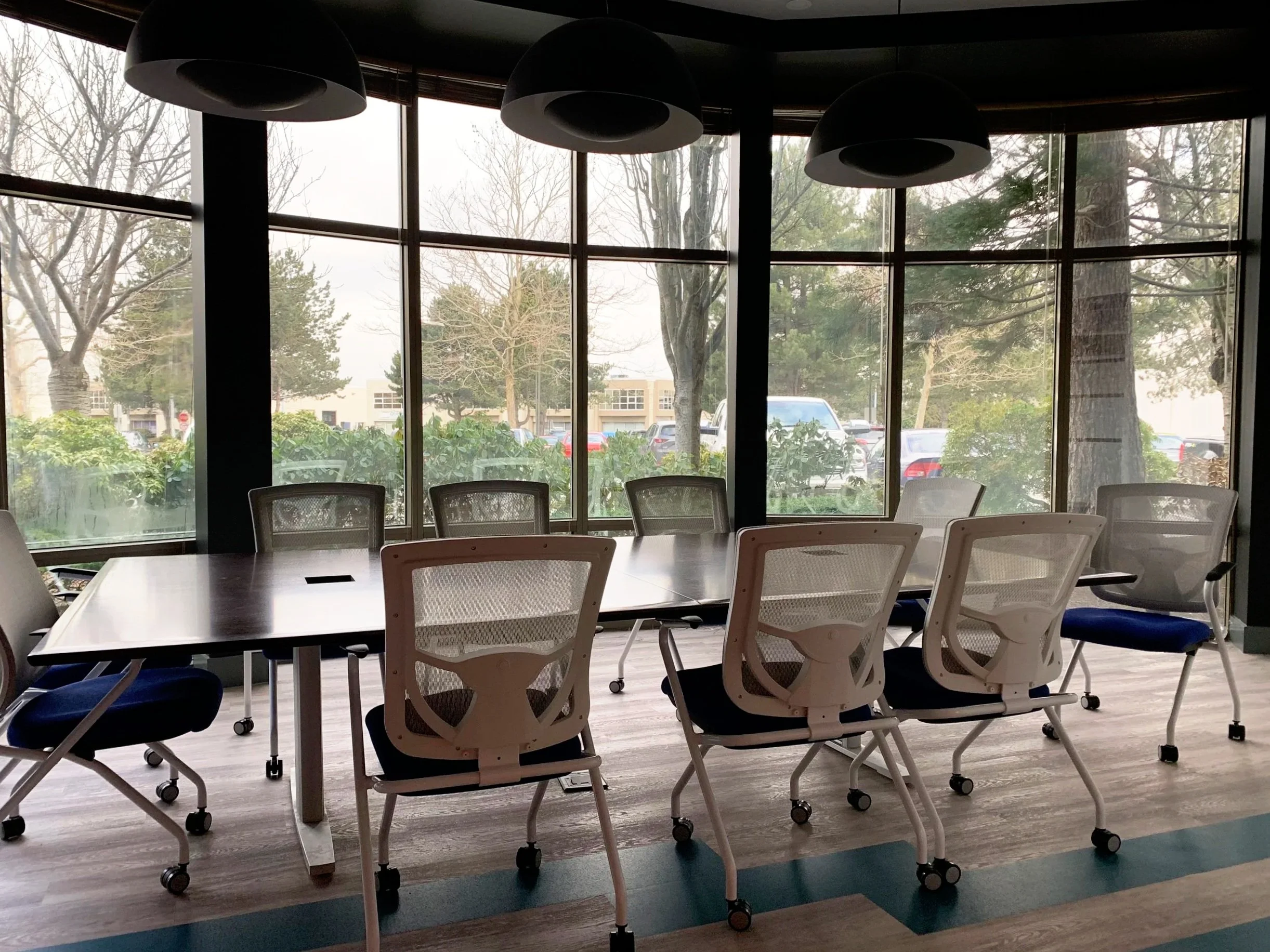 Interior of a conference room with a large rectangular table surrounded by white chairs on wheels, large windows showing trees and parked cars outside, and three black pendant lights hanging from the ceiling.
