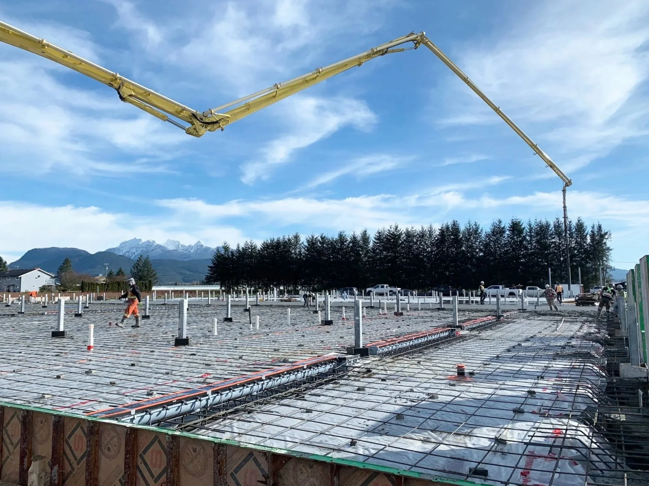 Construction workers working on a building foundation with visible rebar and pipes, with snow-capped mountains and trees in the background.