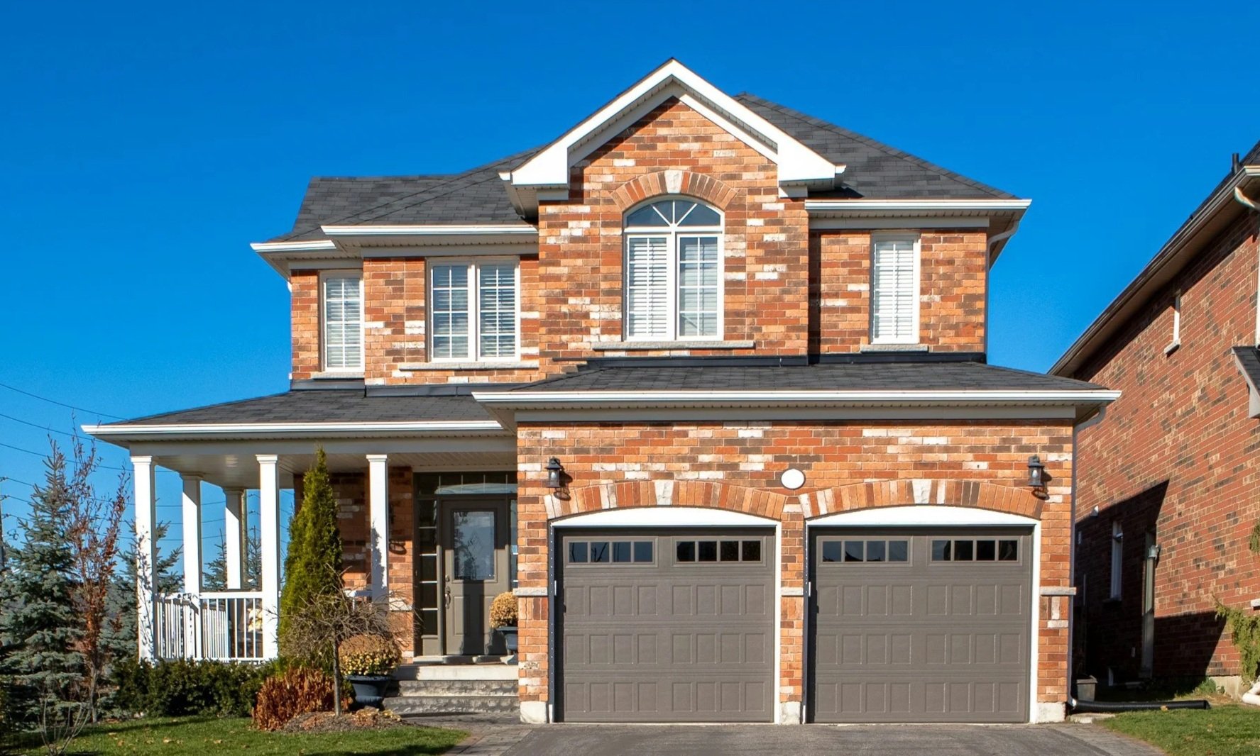 A two-story brick house with a front porch, two attached garage doors, multiple windows with white shutters, and a gable roof under a clear blue sky.